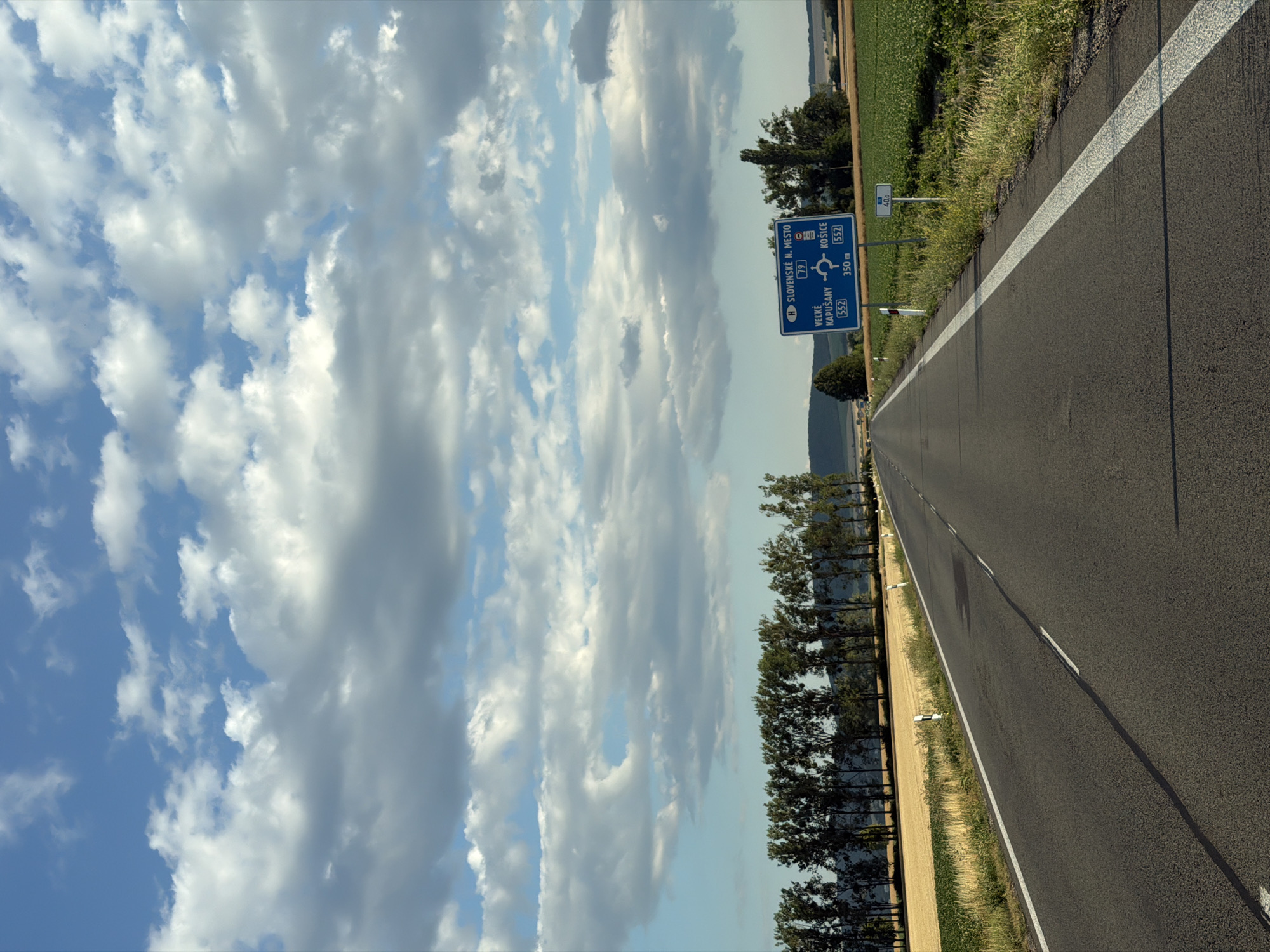 The image shows a straight asphalt road with white lane markings, leading towards the horizon under a sky filled with fluffy white and gray clouds. On the right side of the road, a blue traffic sign is visible, indicating directions to "SLOVENSKE N. MESTO" (Slovenské Nové Mesto), "VEĽKÉ KAPUŠANY 552", and "KOŠICE 552" with a roundabout symbol and distances. The sign also shows "79" which likely refers to a road number. Next to the main sign, a smaller sign indicates "40.0". Fields of yellow and green crops are present on either side of the road, bordered by a line of tall trees in the background. The scene appears to be in a rural area of Slovakia, possibly near Zemplínska Nová Ves, as suggested by the signage. There are no people or vehicles visible in the image.