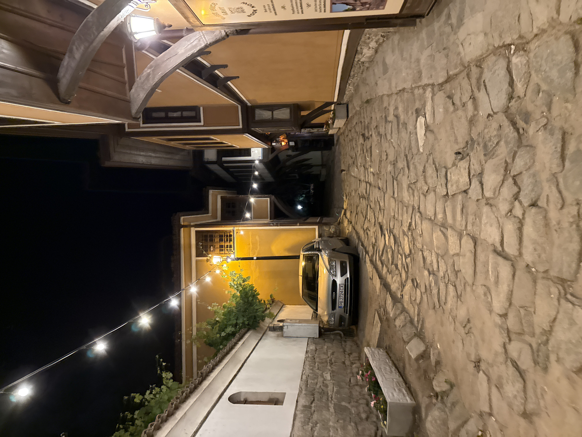 A narrow, cobblestone street in Plovdiv's Old Town is depicted at night. String lights illuminate the scene, casting a warm glow on the traditional Bulgarian Revival architecture. The street is lined with buildings featuring wooden accents and colorful facades, suggesting a historical and charming atmosphere. A silver car is parked on the side of the street, and a planter box with flowers adds a touch of color. A small cat is visible walking down the street, adding a touch of life to the quiet evening. The overall impression is one of a serene and picturesque urban landscape.