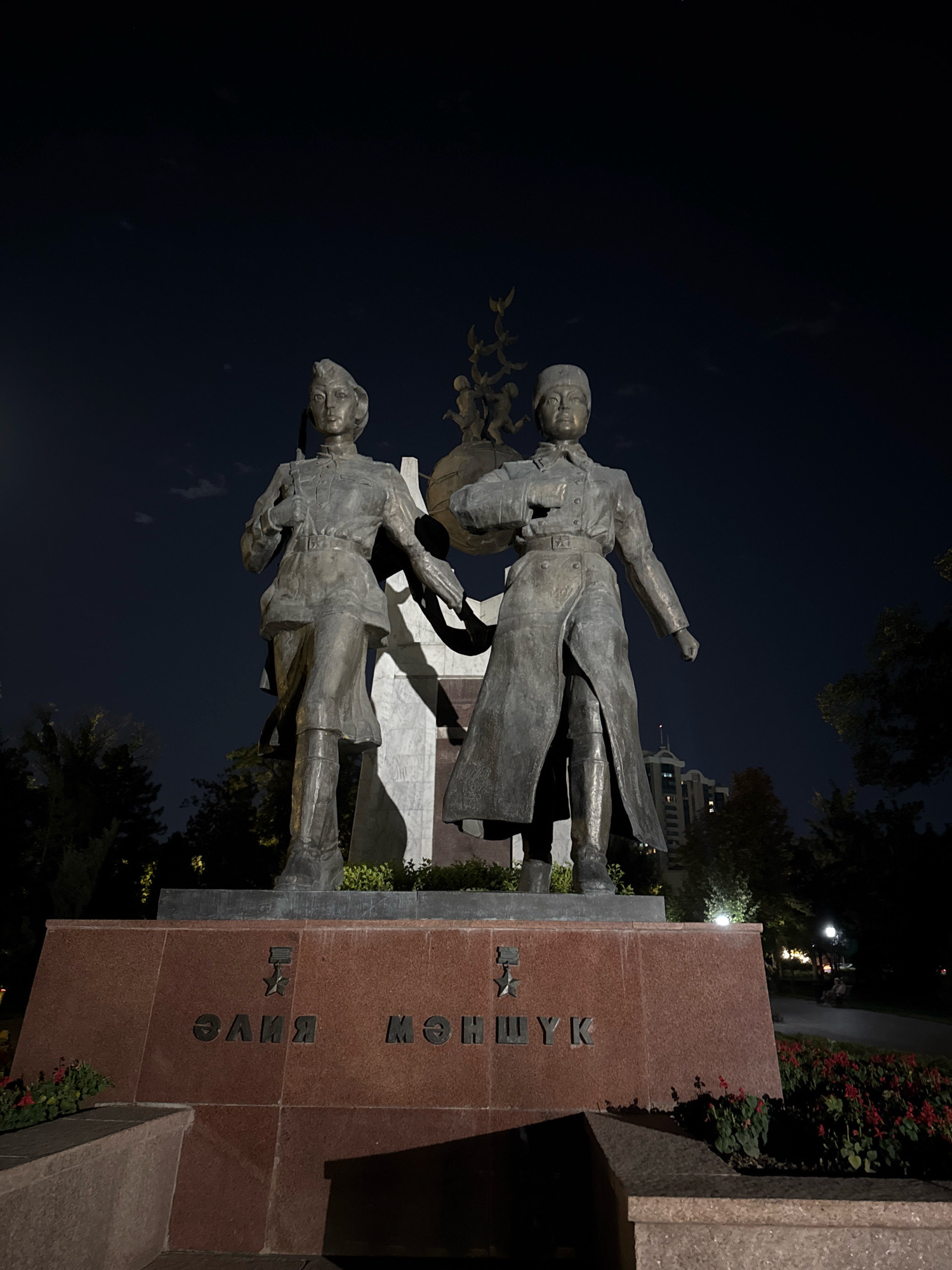 The image depicts a statue at night in Almaty, Kazakhstan. The statue features two female figures, presumably of historical significance, rendered in bronze. They are positioned on a granite pedestal with Cyrillic lettering visible, which reads "Алия Молдагулова" and "Маншук Маметова", indicating the memorial honors two Heroines of the Soviet Union from Kazakhstan. The figures are dressed in military-style uniforms. The statue is illuminated by artificial lights, creating strong shadows and highlighting the textures of the bronze and granite. Behind the statue, trees and a building are dimly visible against the dark night sky. The surrounding area appears to be a park or public square, with some red flowers planted in front of the pedestal. A streetlamp and a distant building with illuminated windows suggest an urban setting.