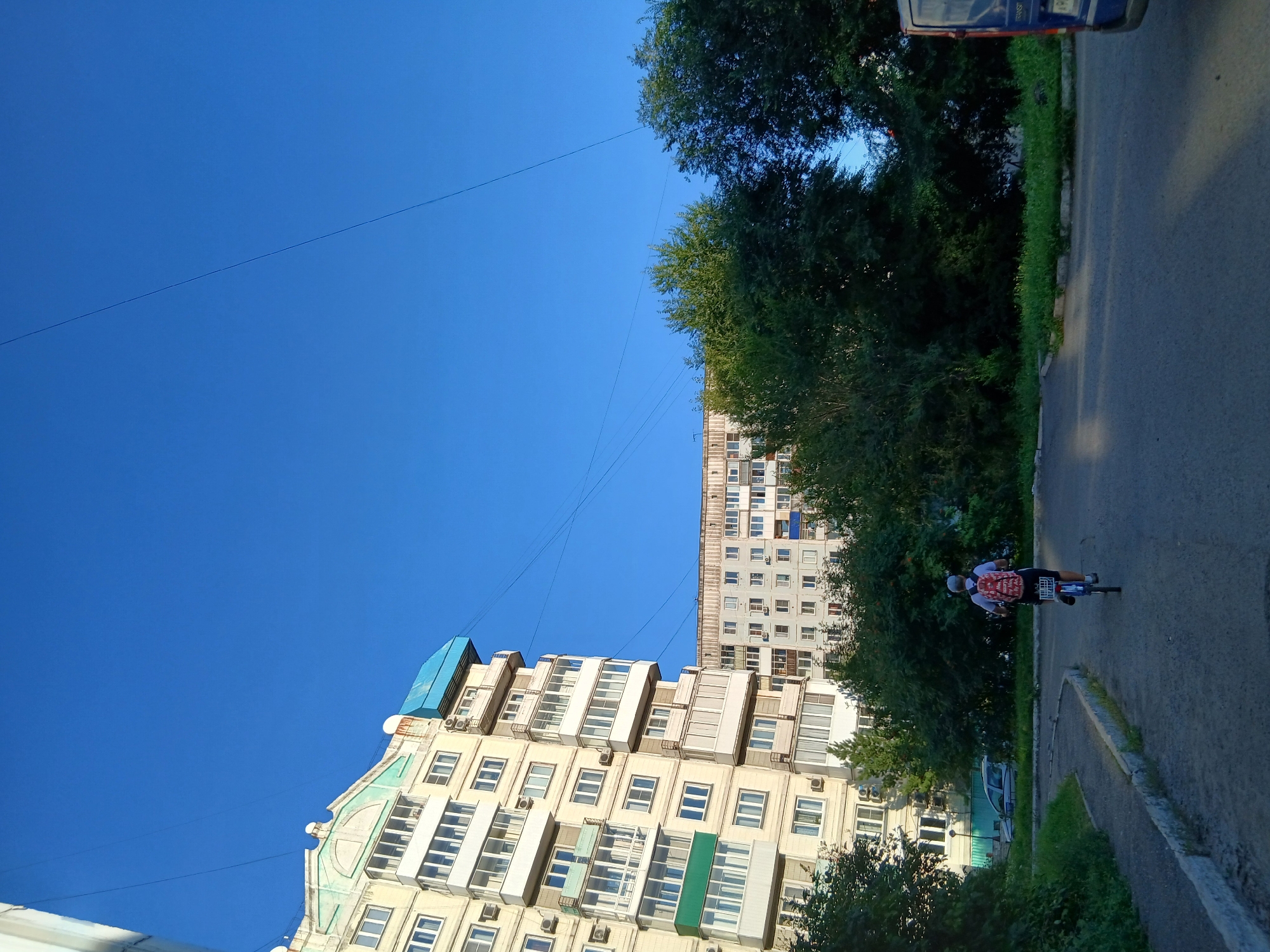 An exterior, eye-level, medium shot captures a clear, bright blue sky over a residential area. In the foreground, a person is seen from behind, riding a bicycle on an asphalt path. The cyclist wears a white shirt and dark shorts, with a red backpack featuring a white pattern. The bicycle has a rear basket. The path is bordered by a concrete curb and a grassy area with some small plants. Behind the cyclist and the grassy area are several mature green trees. Further back, on the left side of the frame, is a tall, light-colored apartment building with many windows and balconies, some with green or blue coverings. To the right of this building, and behind the trees, is another, longer apartment building with a light-colored facade and visible windows. A blue van is partially visible on the right side of the frame. Power lines are visible crisscrossing the sky.