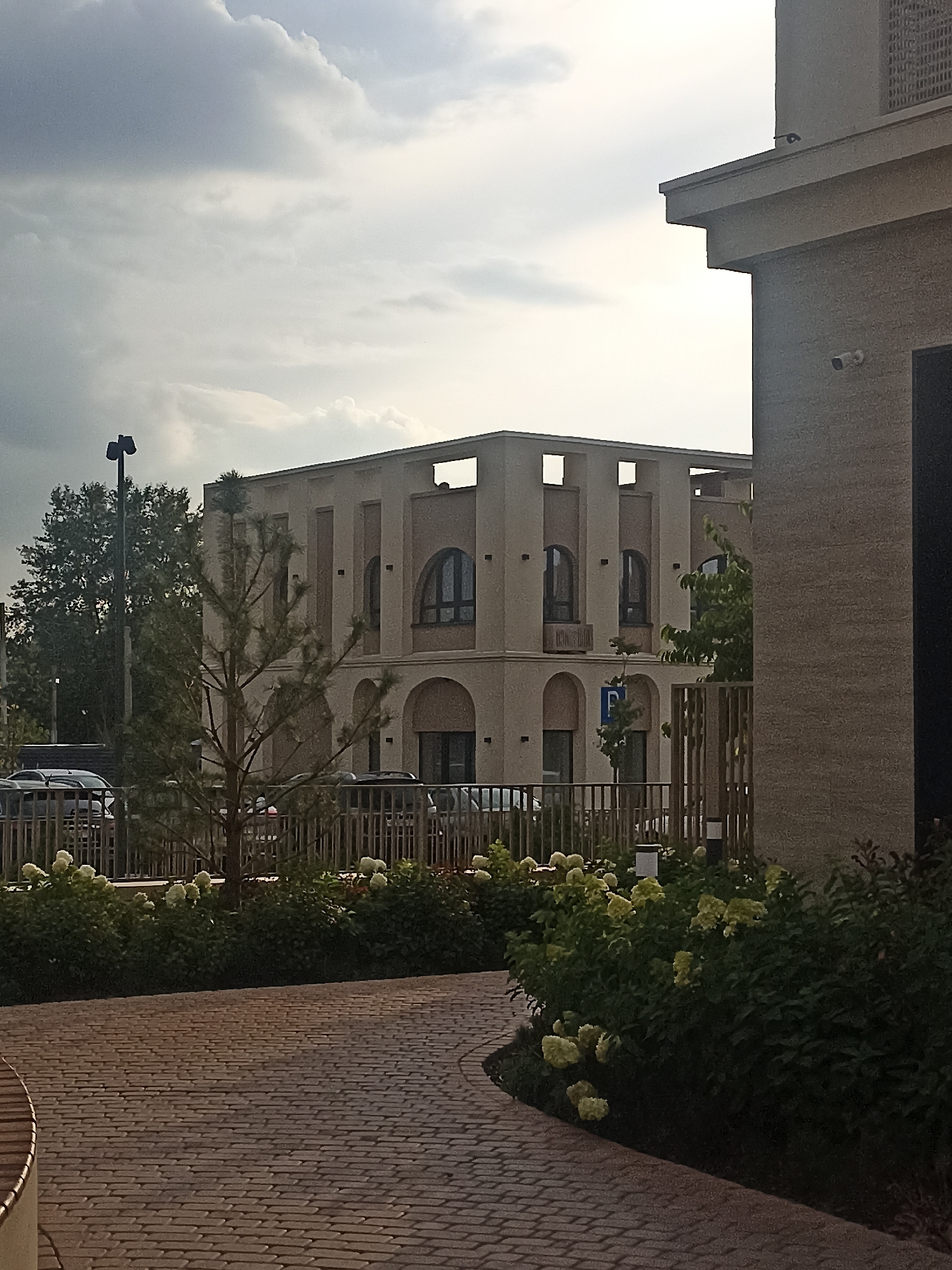 The image depicts an exterior view of a modern building with beige-colored facade, featuring arched windows and recessed decorative elements. In the foreground, a paved walkway curves to the left, made of small, rectangular bricks laid in a pattern. A low, metal fence runs horizontally across the middle ground, behind which several cars are parked. Green bushes and some white-flowered plants are visible along the fence line. A sparse pine tree stands in the center of the frame. In the background, a tall tree and a lamppost are visible to the left of the building. The sky is cloudy with patches of lighter and darker gray. The building appears to be located in a contemporary urban or suburban development. Based on the style of architecture and the presence of a parking sign with a "P" symbol, it is likely a commercial or residential complex. The presence of a parked car and the overall setting are consistent with an outdoor scene in Novosibirsk, Russia.