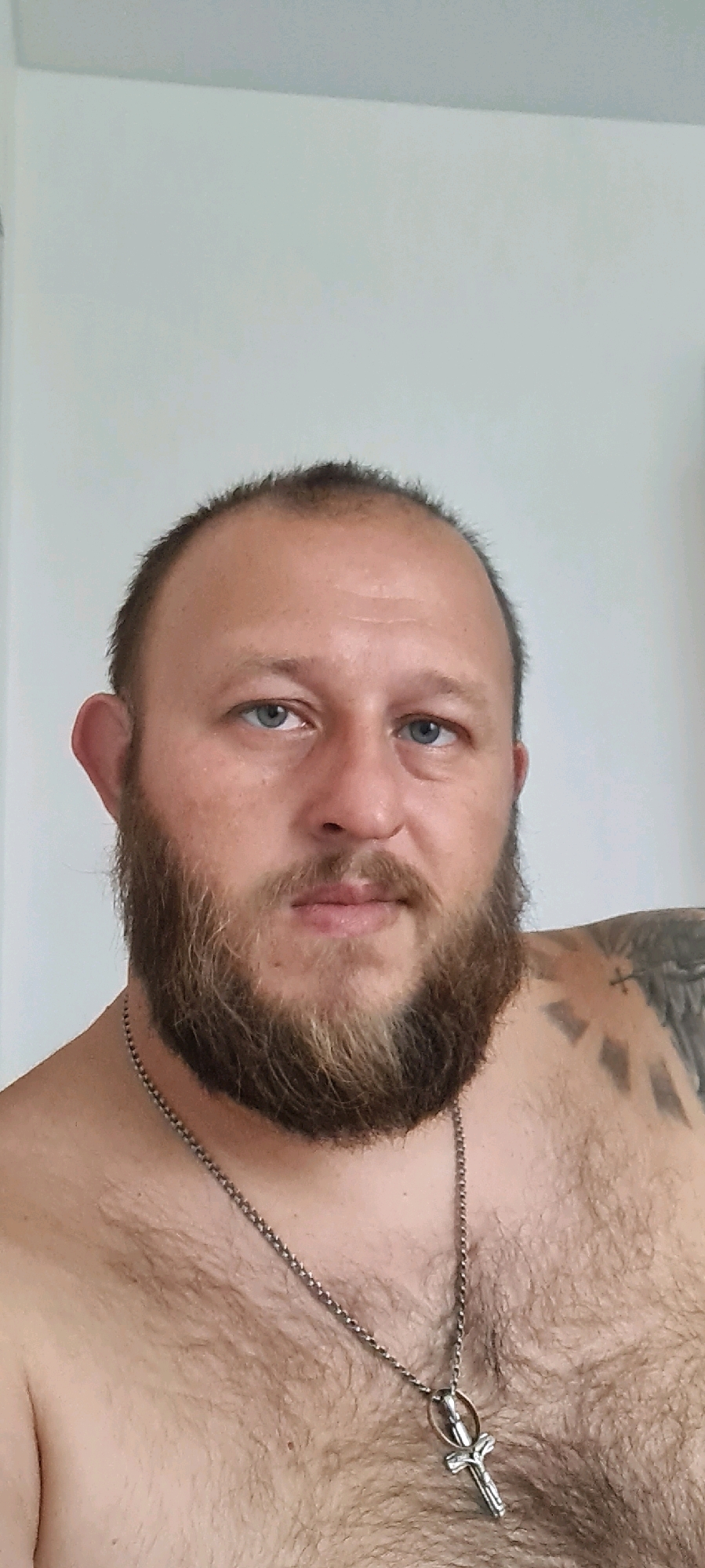 A head and shoulders, close-up, waist-up, medium shot of a Caucasian man, approximately 40 years old, with a full brown beard, a receding hairline, and blue eyes. He is wearing a silver chain necklace with a crucifix pendant. He has a tattoo on his left shoulder that appears to be a cross with radiating lines. The background is a plain white wall. The image was taken indoors.