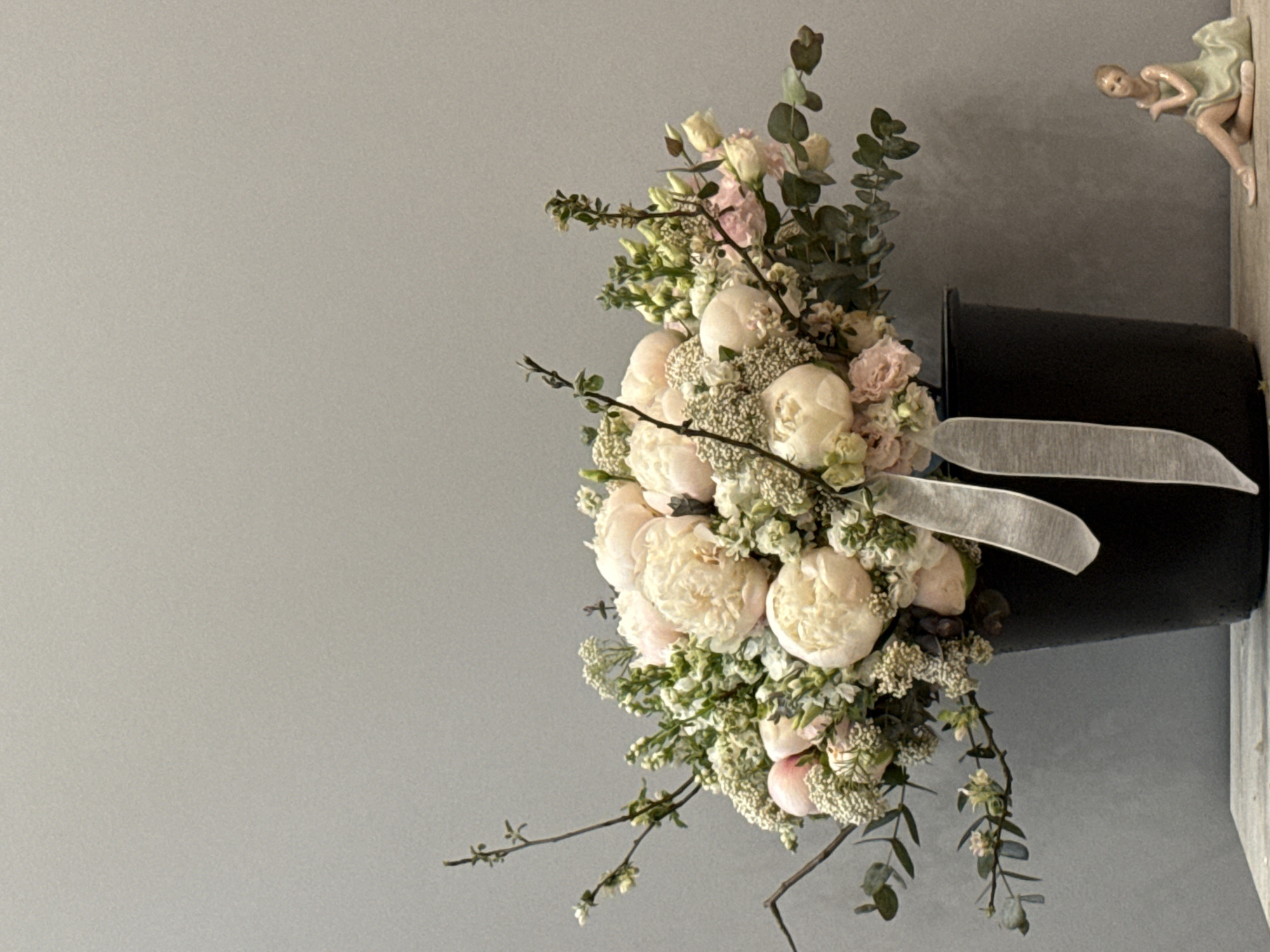The image depicts a bouquet of flowers arranged in a black vase. The flowers are primarily white and pink, consisting of peonies, roses, and greenery such as eucalyptus. A light-colored ribbon is tied around the vase. A ballerina figurine is located on the upper-right corner of the image, positioned on what seems to be a ledge or shelf. The background is a plain gray wall. There is no indication the image was taken in Moscow.