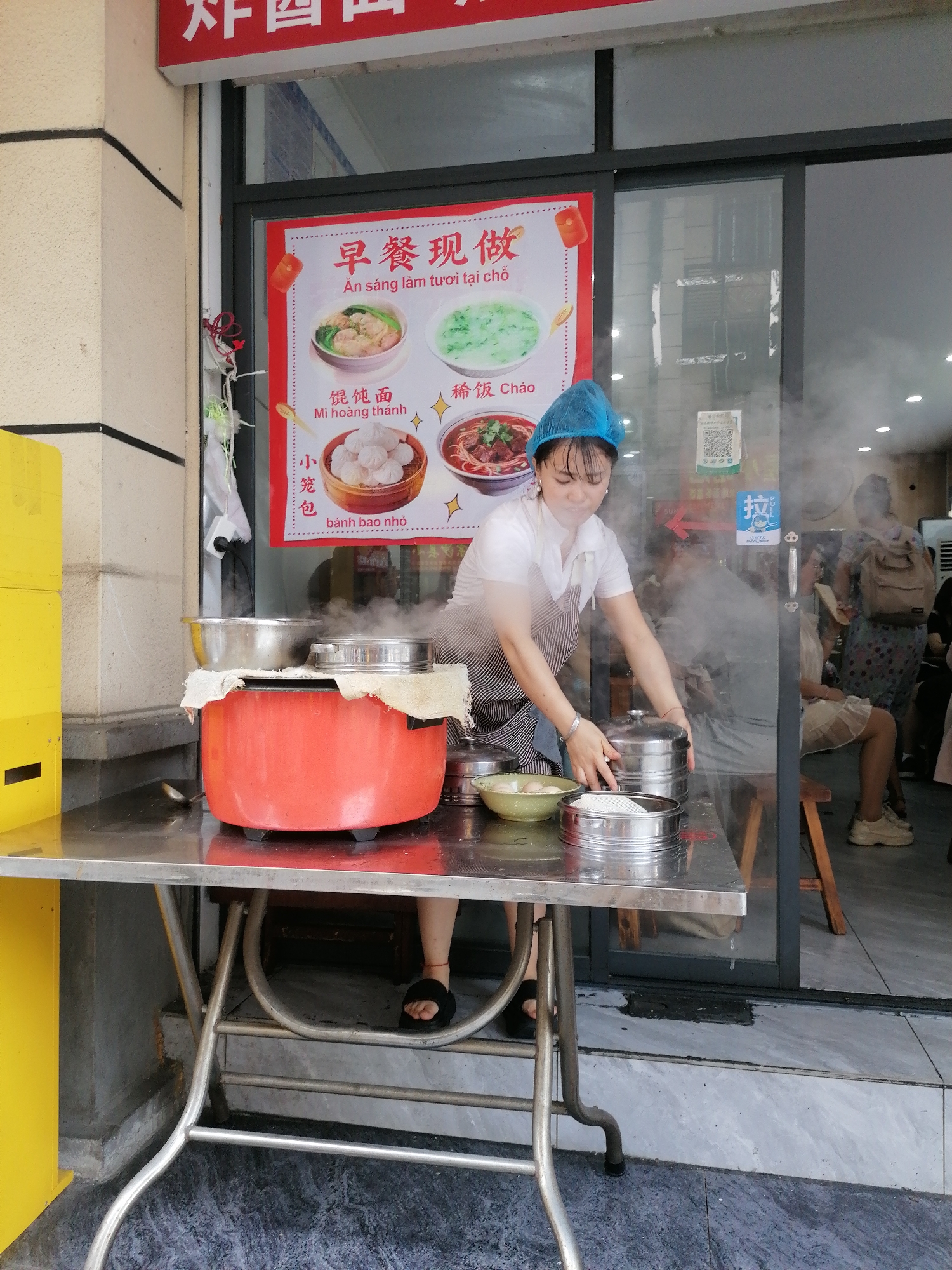 The image shows a woman in Hekou, China, preparing food at an outdoor stand. She is wearing a blue hairnet, a white short-sleeved shirt, and a striped apron. The stand is made of metal and holds a large orange pot, several silver steamer baskets, and a green bowl. Steam rises from the pot and baskets. A poster behind her displays images of various dishes with Chinese and Vietnamese writing. The sign reads, "Breakfast Now Made, An sang lam tu'oi tai cho." The menu items displayed are listed as: "Wonton Noodles - Mi hoang thanh, Tiny Bun - bahn bao nho, Porridge- Chao". In the background, people are seated at tables inside the building. The area appears to be the entrance to a restaurant or food establishment.