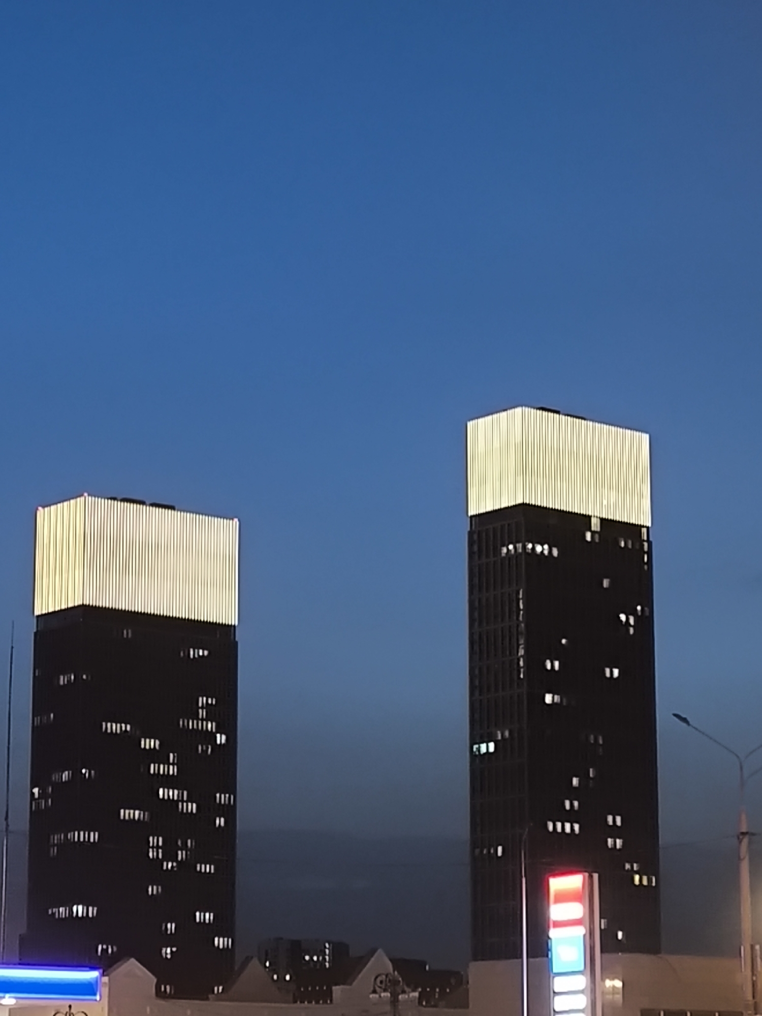 The image shows two tall, dark buildings with illuminated tops in Moscow, Russia. The buildings are rectangular and appear to be towers with windows that are lit. The sky is a deep blue, indicating that the photograph was taken during the evening or nighttime. In the foreground, there are signs and structures that suggest the location is an urban area with commercial establishments.