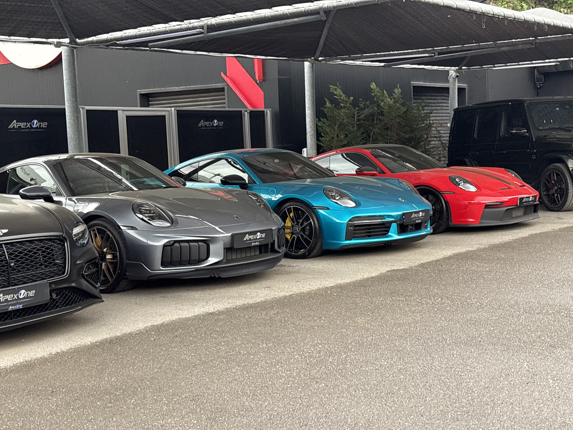 The image shows a lineup of luxury cars parked under a canopy in an outdoor setting. From left to right, there is a Bentley, followed by two Porsche sports cars (gray and blue), another Porsche sports car in red with black stripes, and a black Mercedes-Benz G-Class SUV in the background. "Apex One" logos are visible on the cars and in the background, suggesting the location might be an Apex One luxury car dealership or garage. The location of the city of Sofia, Bulgaria, is unconfirmed in the image.