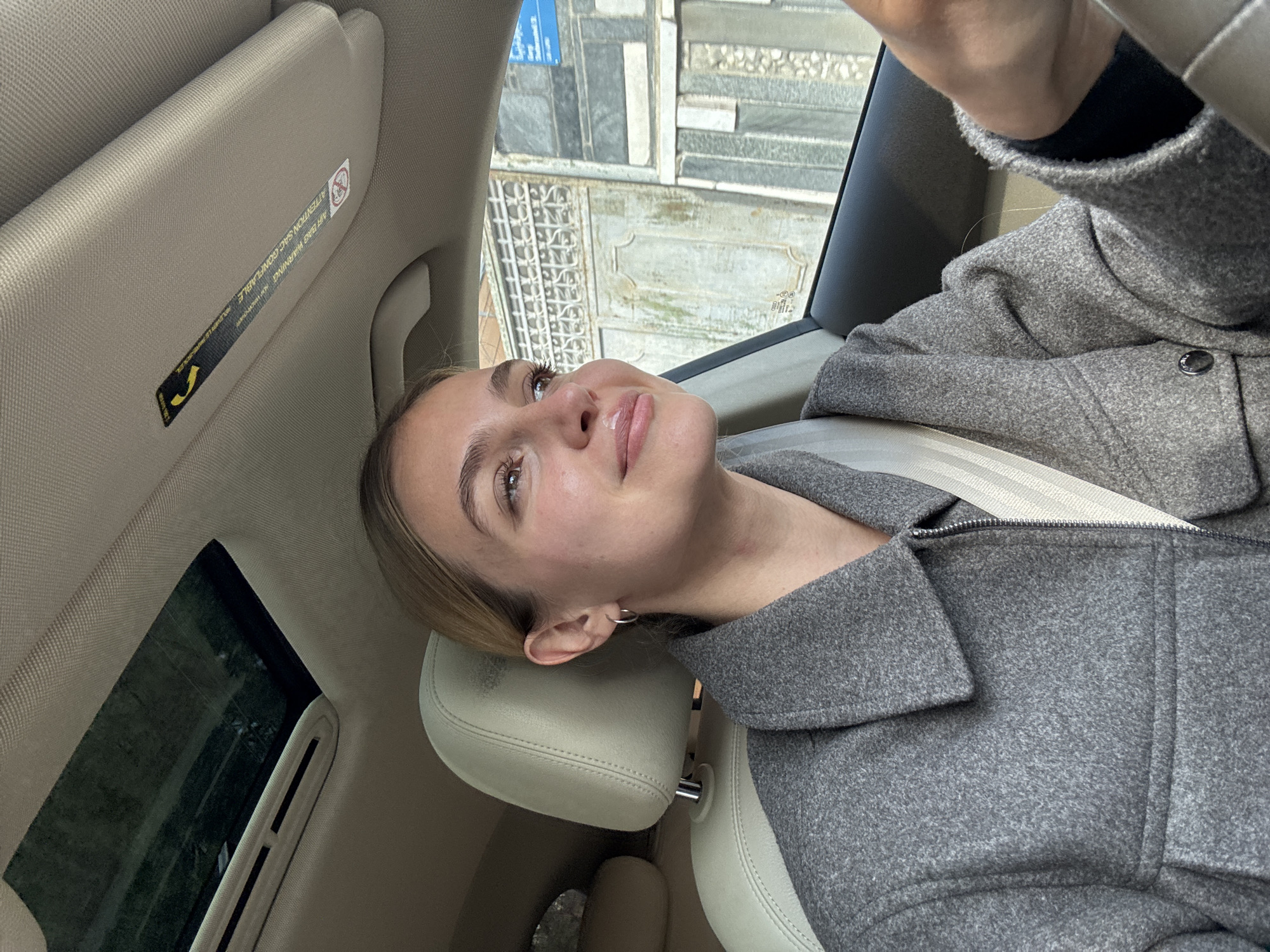 The image depicts a person inside a vehicle, possibly a car, as indicated by the headrest, seatbelt, sun visor, and window visible. The person is wearing a gray coat and appears to be looking upwards. Outside the car window, there is a view of a building with a stone facade. The location in the city is Unknown.