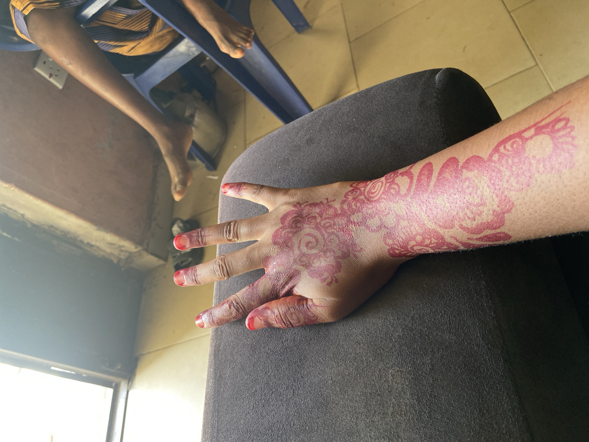A close-up indoor shot reveals a person's hand and arm resting on a gray cushioned surface. The hand and arm are adorned with an intricate red henna-like design that extends from the fingertips up the arm. The fingertips appear to be painted red. The light source comes from a window located on the left side of the frame. In the background, blurred details indicate the legs of a person sitting on a blue chair. The location is Nagazi, Nigeria.