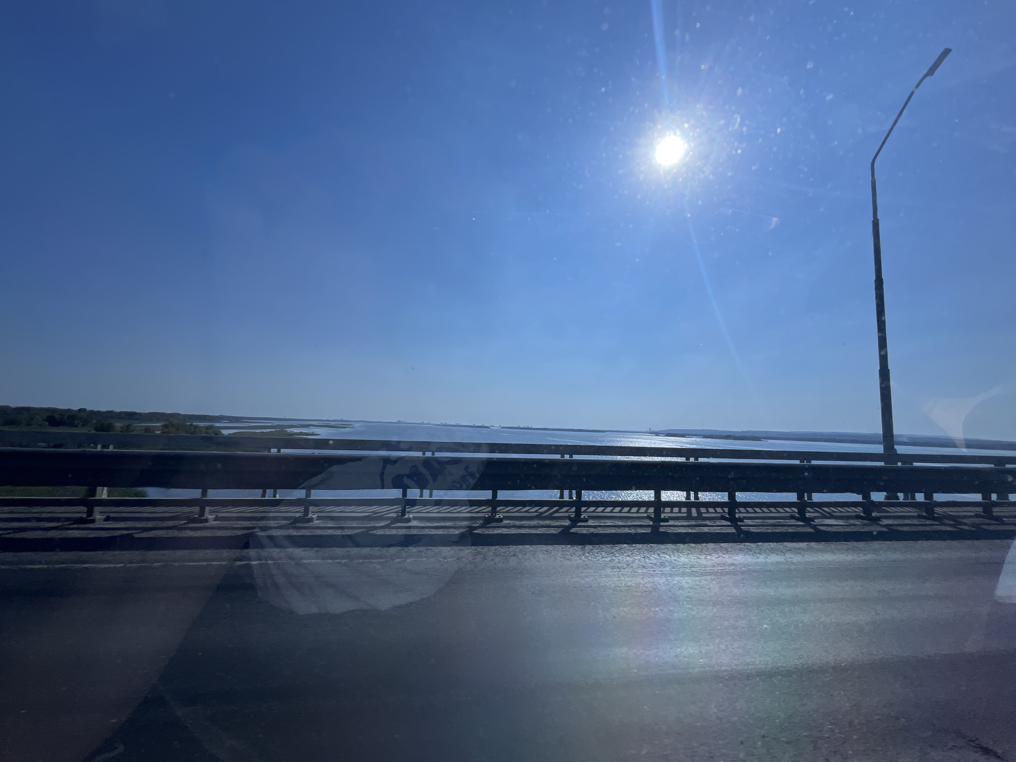This image depicts a view from a bridge over a body of water, likely a river or lake, under a bright blue sky with the sun visible. The sun is positioned in the upper right portion of the frame, causing lens flare and reflections on the camera lens. A tall, dark lamppost stands to the right of the sun. The bridge features a dark metal railing with vertical supports, casting shadows on the asphalt road surface below. In the distance, across the water, a hazy shoreline with vegetation is visible. The overall scene suggests a sunny day in a somewhat rural or suburban setting. The request for the location in "Shumeika, Russia" cannot be verified from the image content alone.