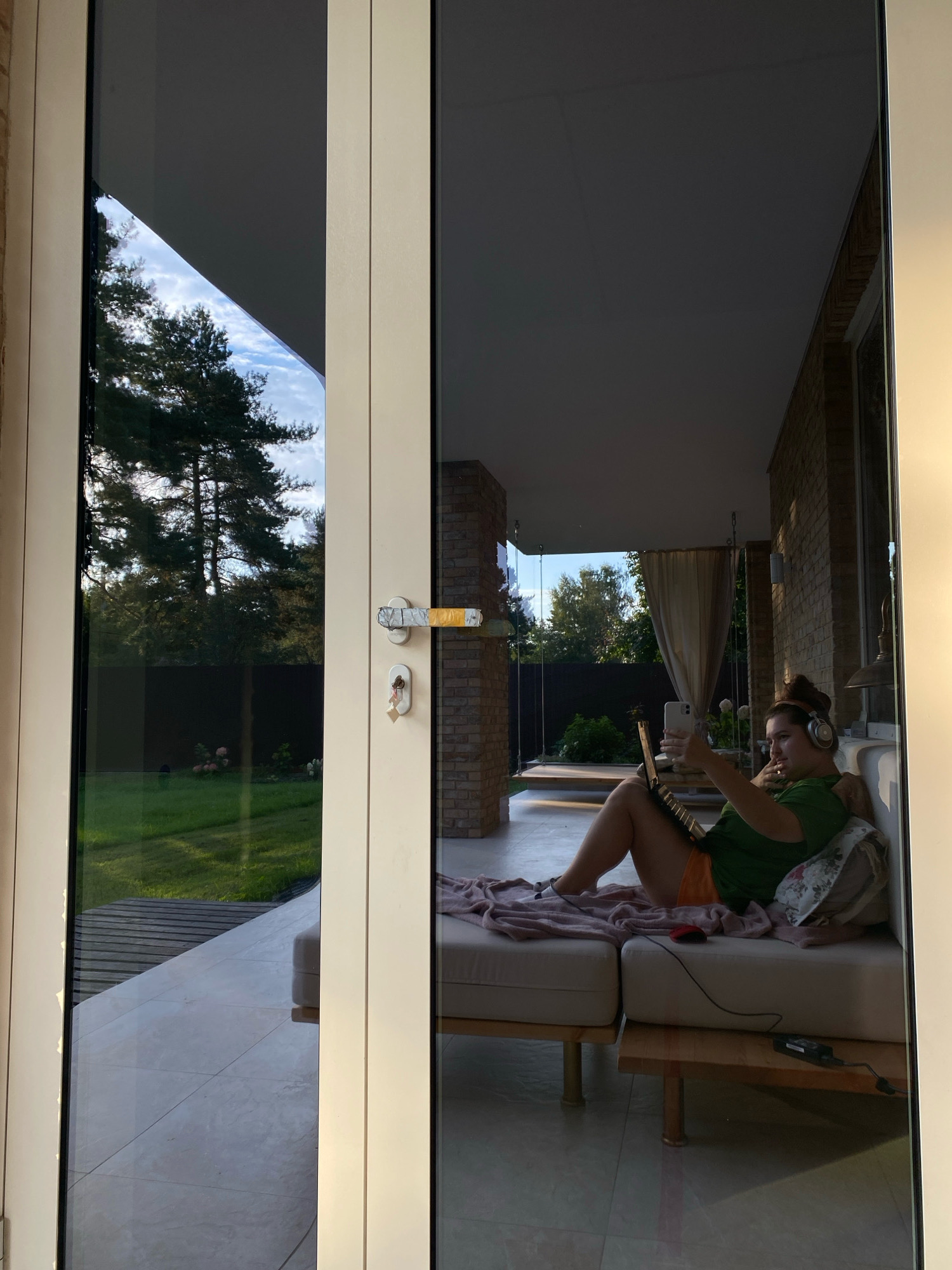 A person is sitting on a couch on a covered patio, looking at a laptop. The person is wearing headphones and holding a phone. The patio is next to a lawn with trees. The scene appears to be in the city of Perm, Russia, based on the architectural style and vegetation. The doors of the patio are closed, and the photographer is outside looking in.