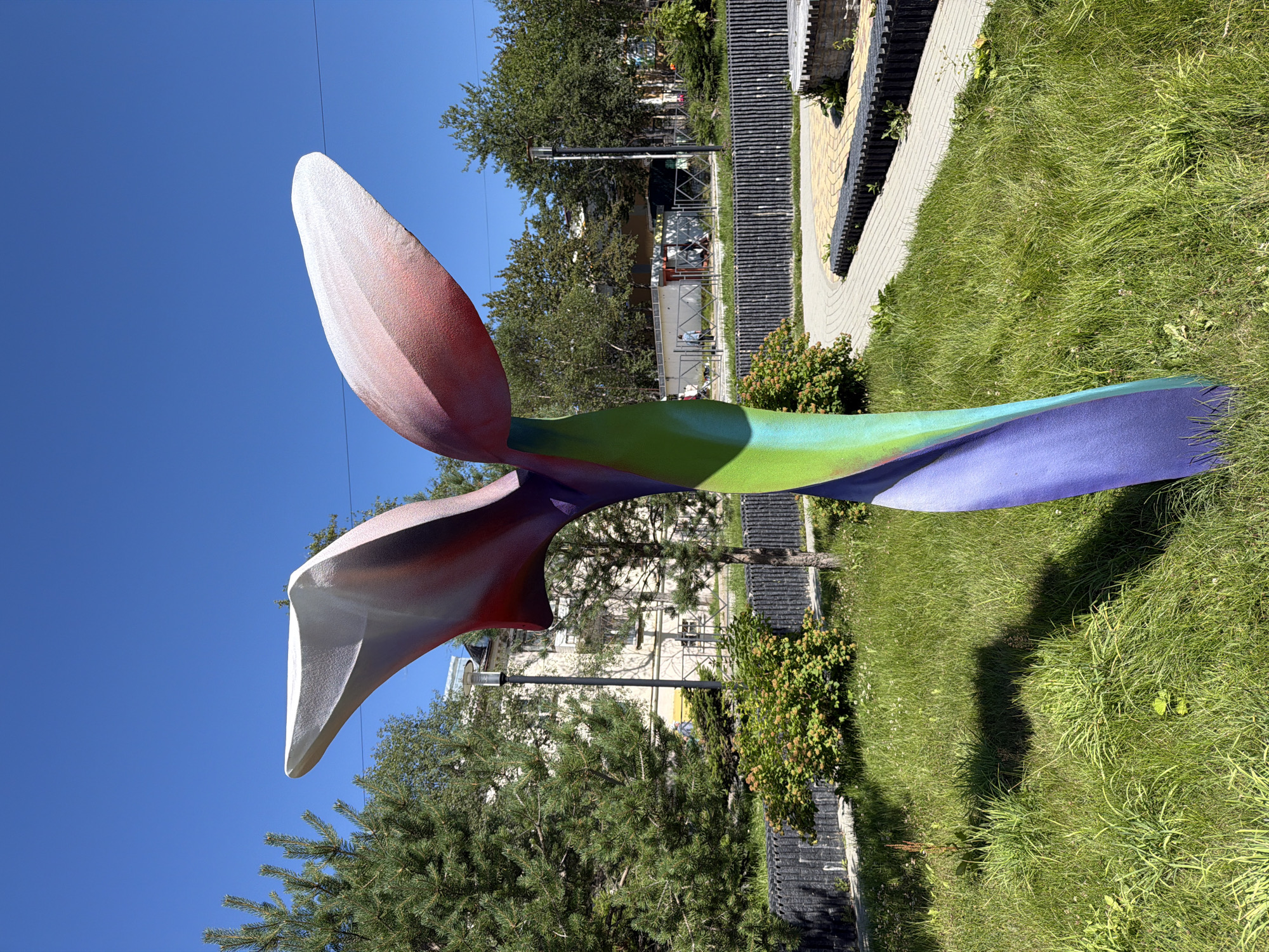 The image shows a colorful, abstract sculpture in a park setting. The sculpture, which appears to be made of metal or a similar material, has a twisted, organic form with petal-like elements at the top. It is painted with a gradient of colors, transitioning from white and pink at the top to red, purple, and green towards the base.
The sculpture is situated on a grassy area with scattered clover. Behind it, a paved walkway curves through the park, bordered by low, dark fencing. Beyond the fence, trees and buildings are visible, indicating an urban environment. In the background, a light-colored building with multiple windows and a streetlamp can be seen. Several trees, including a prominent pine tree to the left of the sculpture, add greenery to the scene. A few people are walking in the distance. The sky is a clear, bright blue.
This scene is located in the city of Okha, Russia, specifically in the area around the Okha Regional Museum of Local Lore. The sculpture is a part of the park that surrounds the museum, likely installed as a public art piece. The overall impression is one of a sunny day in a public space with contemporary art.