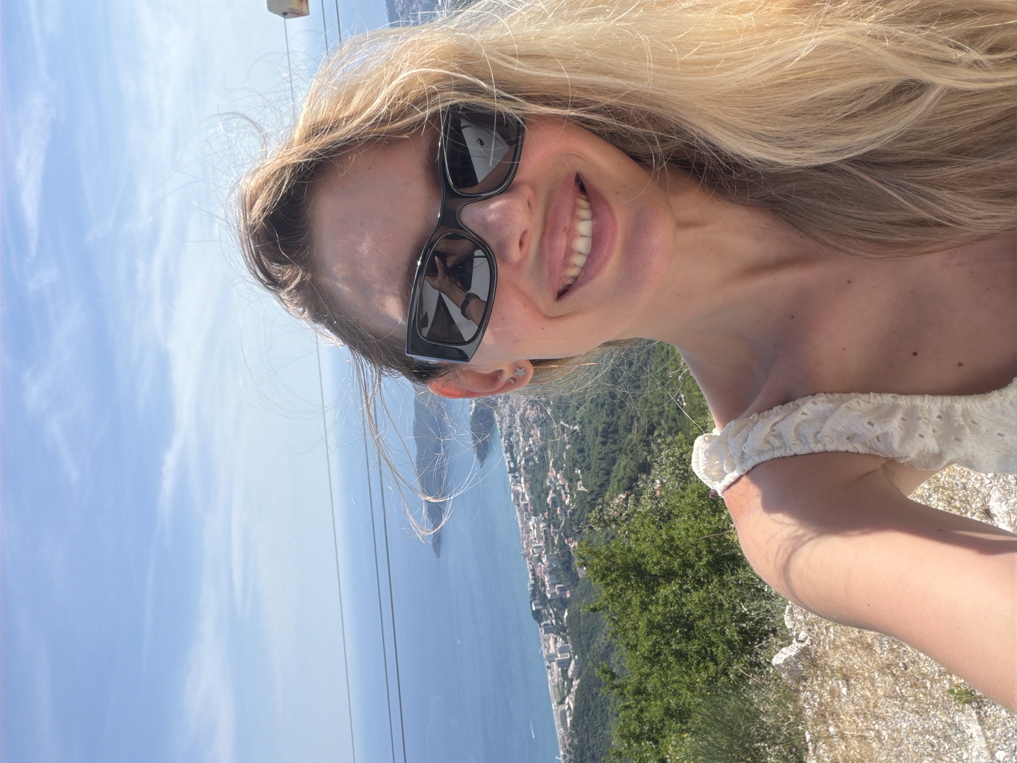 A woman is taking a selfie on a sunny day, with a coastal city and the sea in the background. She has blonde hair and is wearing sunglasses and a white top. The city appears to be Boreti, Montenegro, with buildings clustered along the coastline and hills covered in greenery behind it. The sea is a vibrant blue, and the sky is clear with a few wispy clouds. The woman is smiling and looking at the camera. Her left ear has a star-shaped earring. In the reflection of her sunglasses, another person's arm and hand holding a phone or camera can be seen. There are two parallel wires stretching across the upper part of the image, possibly part of a cable car system.
