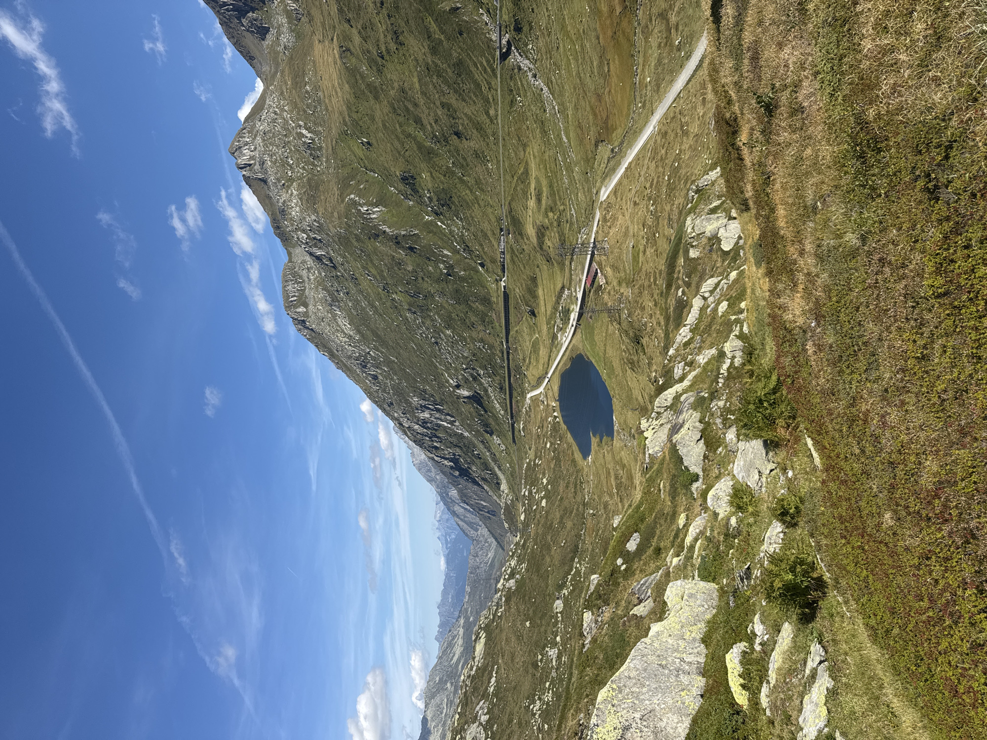 The image depicts a mountainous landscape under a bright blue sky with scattered clouds. In the foreground, a grassy slope is covered with low-lying vegetation and rocks. A winding road and a small, dark blue lake are visible in the middle ground. Beyond the lake, a series of mountains rise, their slopes covered in grass and rock. In the distance, a valley stretches out, with more mountains forming the horizon. The scene appears to be a natural outdoor environment, possibly in a high-altitude region. The area is characterized by rugged terrain, open skies, and sparse vegetation. This is the Gotthard Pass, a high Alpine pass in Switzerland, located near the town of Airolo.