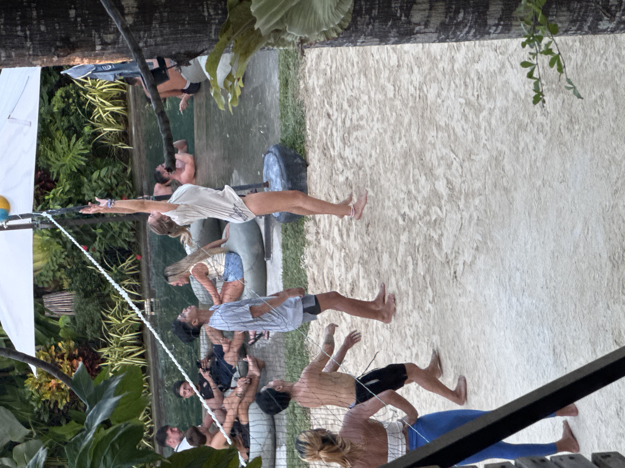 The image captures a vibrant outdoor leisure scene at a tropical resort in General Luna, Philippines. The primary setting is a sandy court, likely for beach volleyball, bordered by lush green foliage and a clear swimming pool or shallow water feature. A white rope net stretches across the court, indicating an ongoing or imminent game.
Numerous individuals are visible throughout the frame, engaged in various recreational activities. On the left side of the net, a woman in a light-colored top and shorts stands barefoot on the sand, her arms extended upwards as if playing volleyball or stretching. She wears a subtle ankle bracelet. Behind her, several people are seen in or around the water, some appearing to relax on a submerged ledge, including a man with visible tattoos. A yellow and blue ball is partially visible near the net on this side.
On the right side of the net, several other participants are on the sandy court. A man in a light grey t-shirt and dark shorts stands barefoot, while a shirtless man in black shorts looks towards the net, his back partially to the viewer. Another person in blue leggings is also visible on the sand further back. More individuals are scattered around the periphery, some watching, some conversing, and others partially obscured by the net or foliage.
The environment is rich with tropical plants, wooden structural elements, and white canvas awnings, suggesting a comfortable and shaded resort amenity. The lighting indicates it is daytime, with a soft, ambient glow. The overall atmosphere is relaxed and social, reflecting a leisurely outdoor gathering. No discernible text is visible within the image.