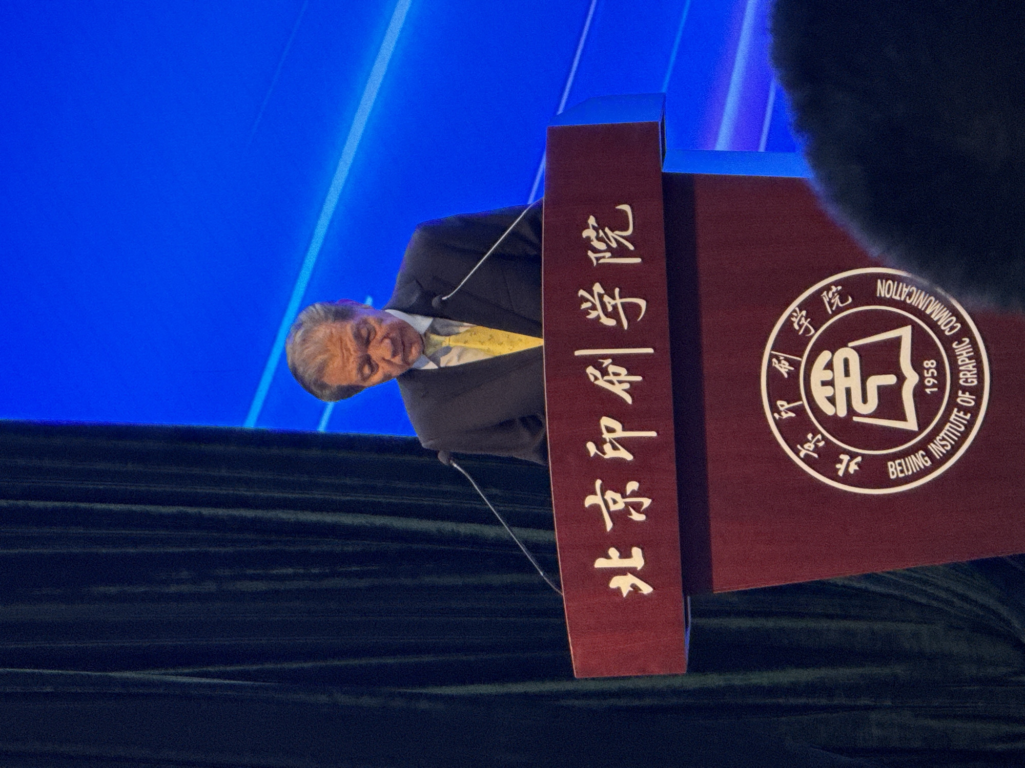 The image shows a man in a dark suit and yellow tie speaking at a podium. The podium is dark red with white text and a circular emblem. The emblem contains Chinese characters and English text that reads "BEIJING INSTITUTE OF GRAPHIC COMMUNICATION 1958". The man is looking down and appears to be reading from notes. Behind him, a dark green curtain is visible on the left, and a bright blue background with diagonal lines is on the right. The scene is indoors, likely at an event or conference. The location is specified as Daxing District, China.