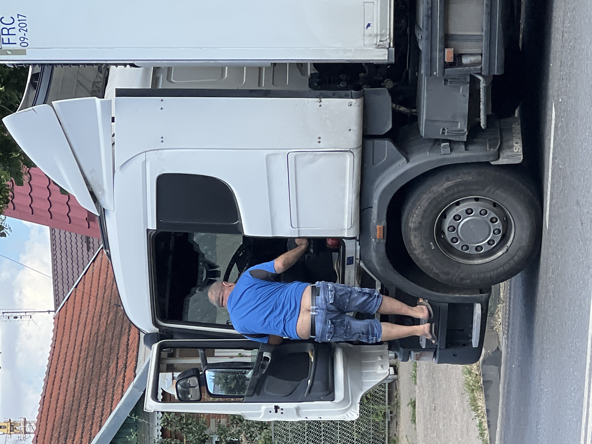A man wearing a blue shirt and denim shorts is climbing into the cab of a white semi-truck. The truck's door is open, revealing the interior. The man is barefoot, wearing flip-flops on his feet as he steps onto the truck's running board. In the background, there are red-tiled roofs of houses and trees. The scene appears to be on a street in Krasnodar, Russia, as indicated by the "FRC 09-2017" marking on the truck, which is a common vehicle registration format in Russia.