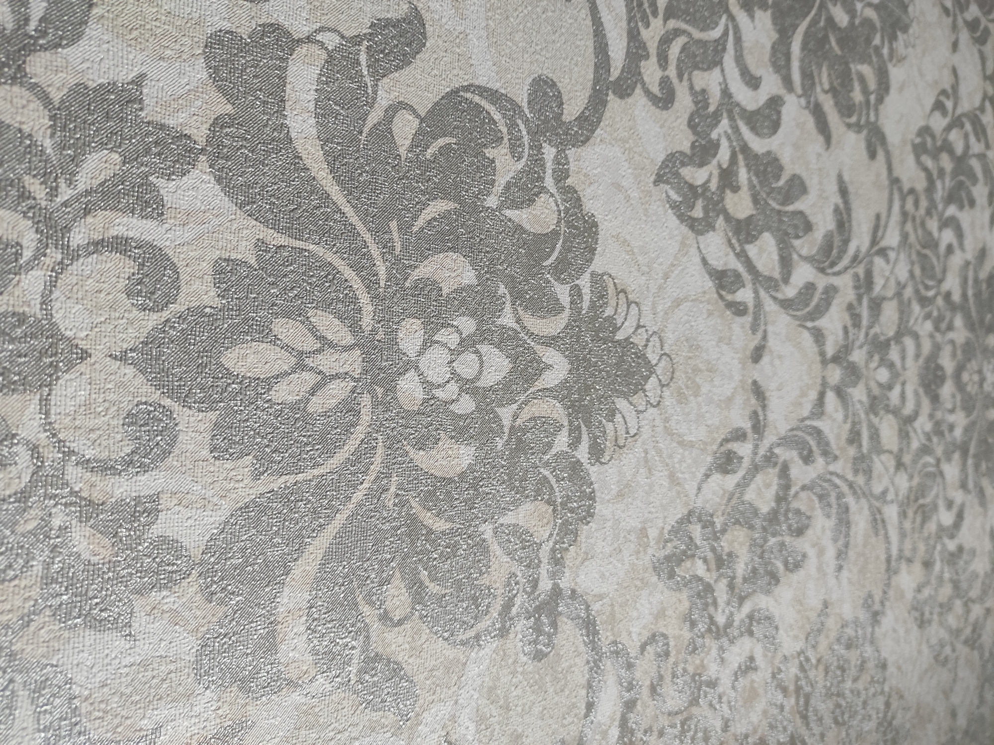 The image displays a close-up view of textured wallpaper with a damask pattern. The wallpaper has a light beige background and features a repeating ornate floral motif in a darker gray, textured finish. The pattern is characterized by swirling vines, leaves, and flower-like elements. The perspective is angled, with the foreground showing more detail of the texture and pattern, while the background appears slightly out of focus. There are no people, actions, or specific environmental elements visible, as the image solely focuses on the surface pattern of the wallpaper. There is no information in the image that can be used to determine its location in the city of Achinsk, Russia.