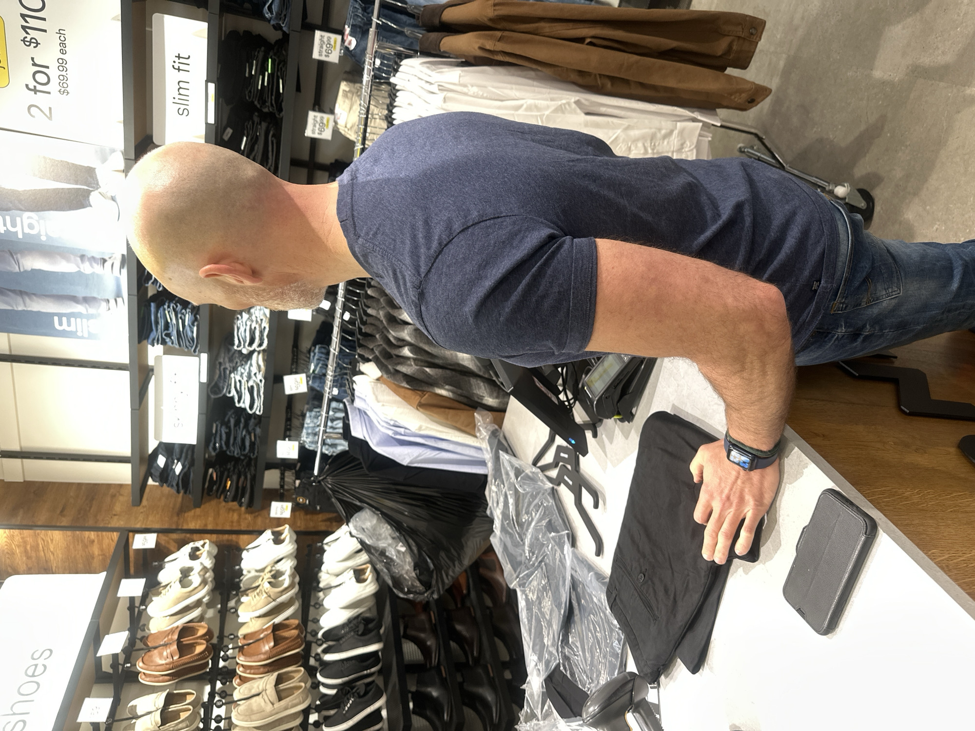 A bald Caucasian male, appearing to be in his late 40s or early 50s, stands at a retail counter in a clothing store. He is wearing a dark blue t-shirt and blue jeans. On his left wrist, he wears a smartwatch. He is facing away from the camera, looking towards the racks of clothing behind the counter. On the counter, a black wallet-sized object, a folded pair of black pants, and clear plastic packaging are visible. A retail scanner is positioned to the left of the wallet object, and a point-of-sale device is to the right. The background reveals shelves stocked with men's shoes and clothing, along with promotional signage. The environment is a well-lit retail store. The provided context suggests this image is from Coffs Harbour, Australia, but no specific landmarks or indicators within the image confirm this location.