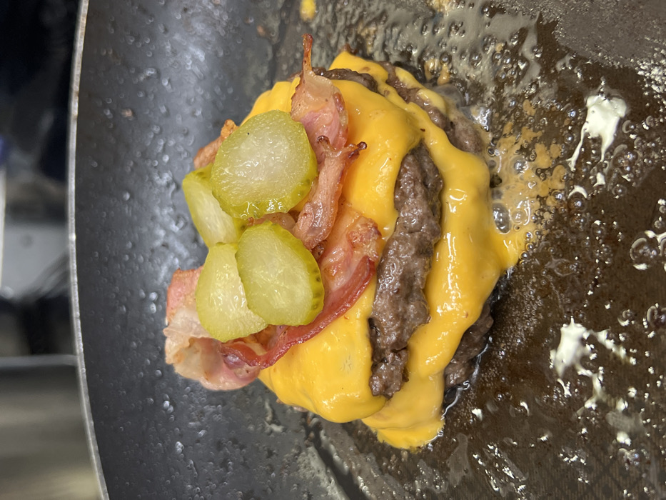 A close-up, slightly overhead view displays a double-stacked burger assembly within a dark frying pan or griddle, located in Budva, Montenegro. The assembly features two browned meat patties, each topped with a melted slice of yellow American-style cheese. Several strips of cooked bacon, varying in crispness, are layered above the uppermost cheese. On top of the bacon, multiple light green, circular slices of dill pickles are arranged. The base of the burger is surrounded by a pool of bubbling grease or cooking oil within the pan. The background is out of focus, indicating an indoor kitchen environment. No individuals are visible.