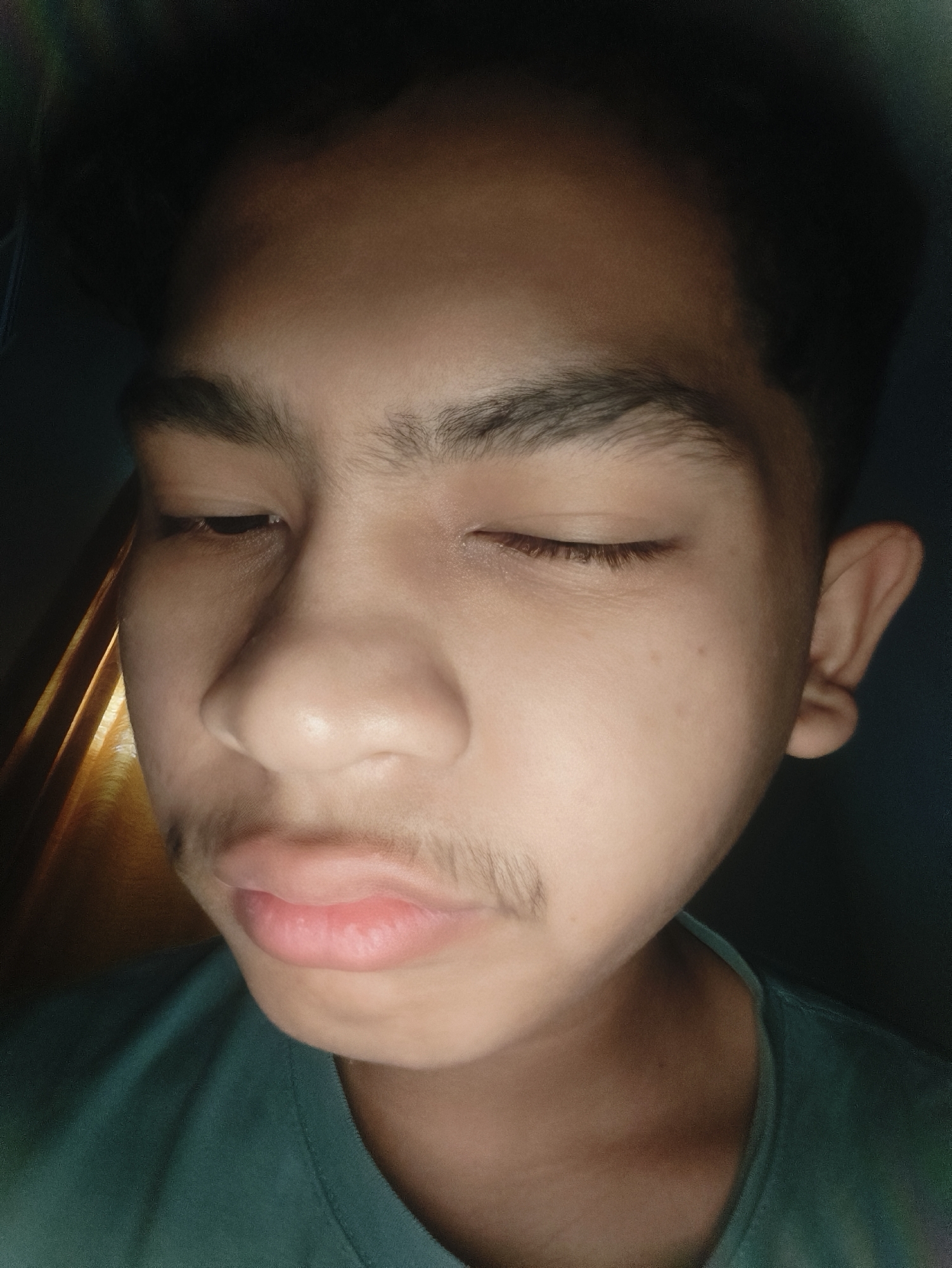 This image, captured in Lhokseumawe, Indonesia, features a close-up portrait of a single individual, a young male with light brown skin, positioned at a slight angle. The subject's face occupies the majority of the frame, with both eyes closed. Dark, thick eyebrows are visible, along with eyelashes on the closed eyelids. Sparse dark facial hair is present on the upper lip and jawline. The subject is wearing a dark teal or green crew-neck t-shirt, partially visible around the neck and shoulder area. The lighting is uneven, with a warm, yellowish light source illuminating the left side of the subject's face (viewer's right) and creating a shadow across the right side of the face. The background is largely dark and indistinct, with a vertical strip of diffused light visible on the far left, suggesting an indoor environment. No other people, subjects, or direct interactions are depicted.