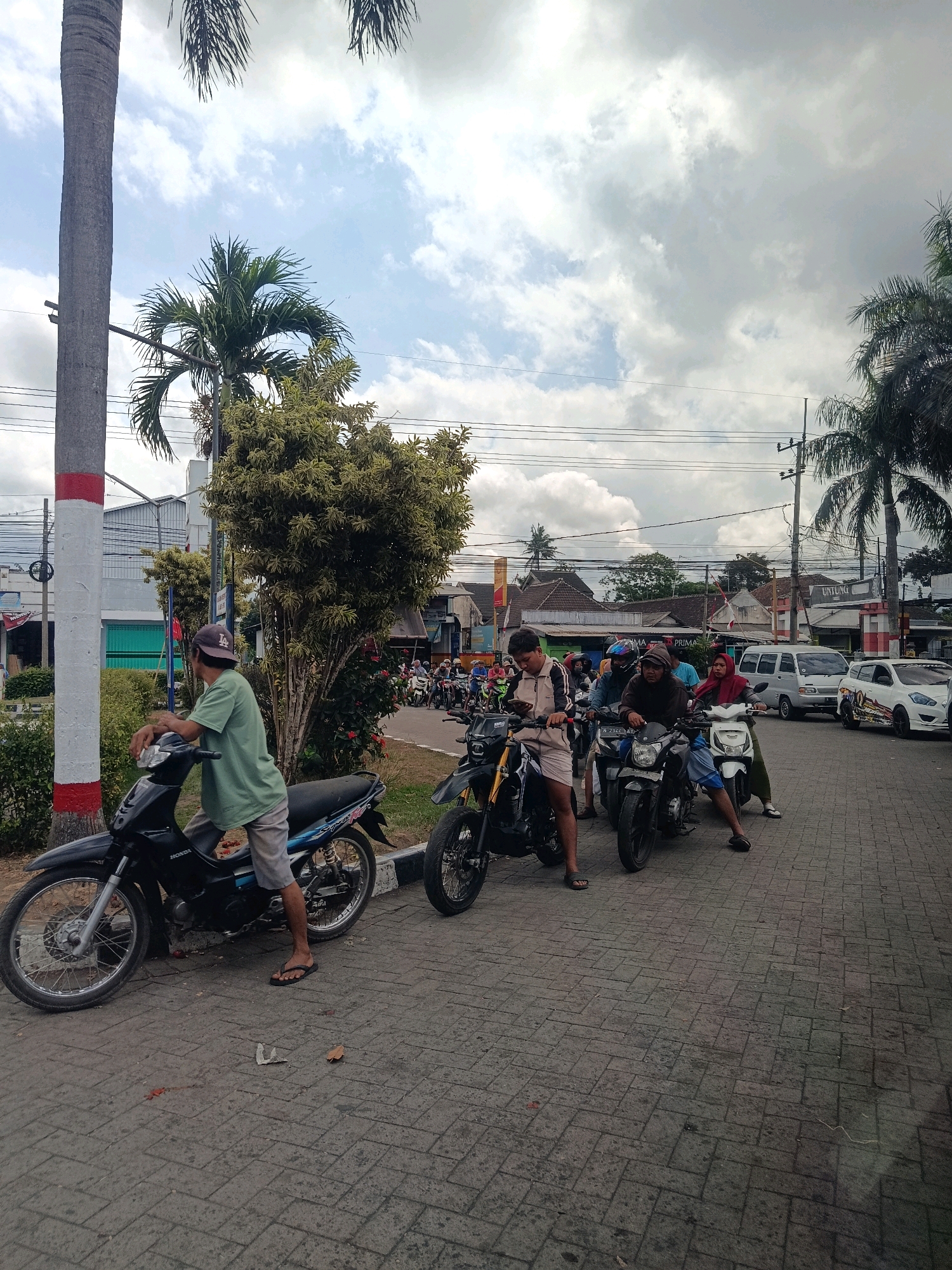 A wide shot captures a street scene in Tempeh Lor, Indonesia, under a cloudy sky. A man in a green t-shirt and gray shorts sits on a black Honda motorcycle, facing left. He is wearing a dark baseball cap. To the right, a line of motorcycles and their riders are stopped, facing right, suggesting a traffic pause or intersection. Among the riders are several individuals, some wearing helmets and others with headscarves. One person is on a white scooter. Behind the motorcycles, a silver van and a white car are visible. Buildings with tiled roofs line the street, and palm trees and other foliage are present. The ground is paved with textured gray bricks. Several small red and orange items are scattered on the pavement in the foreground, along with a crumpled piece of paper.