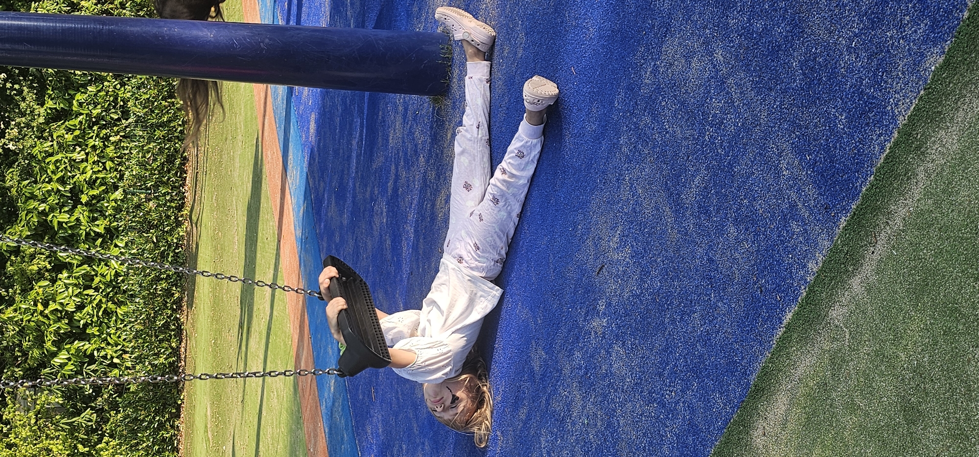 A young girl with blonde hair, wearing white pajamas and pink clogs, lies on her back on a blue rubber playground surface, holding onto a black swing seat. The swing is suspended by chains from above. The playground is bordered by green turf. In the background, a dense green hedge forms a backdrop. A large blue pole is visible on the right side of the image. The scene is set outdoors during the day, likely at a playground in Groenlo, Netherlands, given the context.