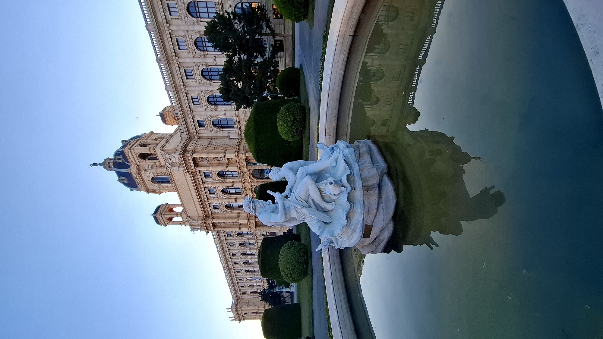 The image depicts a statue in a fountain in front of the Kunsthistorisches Museum (Museum of Fine Arts) in Vienna, Austria. The sky is a clear, pale blue, indicating either dawn or dusk. The museum, an imposing Baroque building with a large central dome and ornate architectural details, occupies the background. In the foreground, a white marble sculpture of two figures, one adult and one child, is situated on a rocky base within a circular fountain. The water in the fountain is calm, reflecting the museum building and the sculpture. The surrounding area includes manicured green lawns and neatly trimmed hedges. The scene is serene and bathed in soft, natural light.