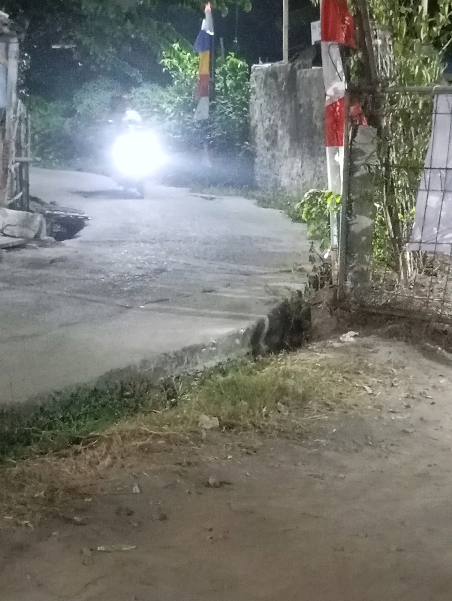A motorcycle with its bright headlight on is moving on a paved road at night. The motorcycle is a blur of light, making it difficult to discern details. To the right of the road, there is a wall and some green foliage. A flag with red and white stripes is visible near the foliage. The road appears wet, possibly due to recent rain, and there are patches of grass and dirt along the roadside. The scene is dimly lit, suggesting it's nighttime in an outdoor urban or semi-urban environment in Babelan, Indonesia.