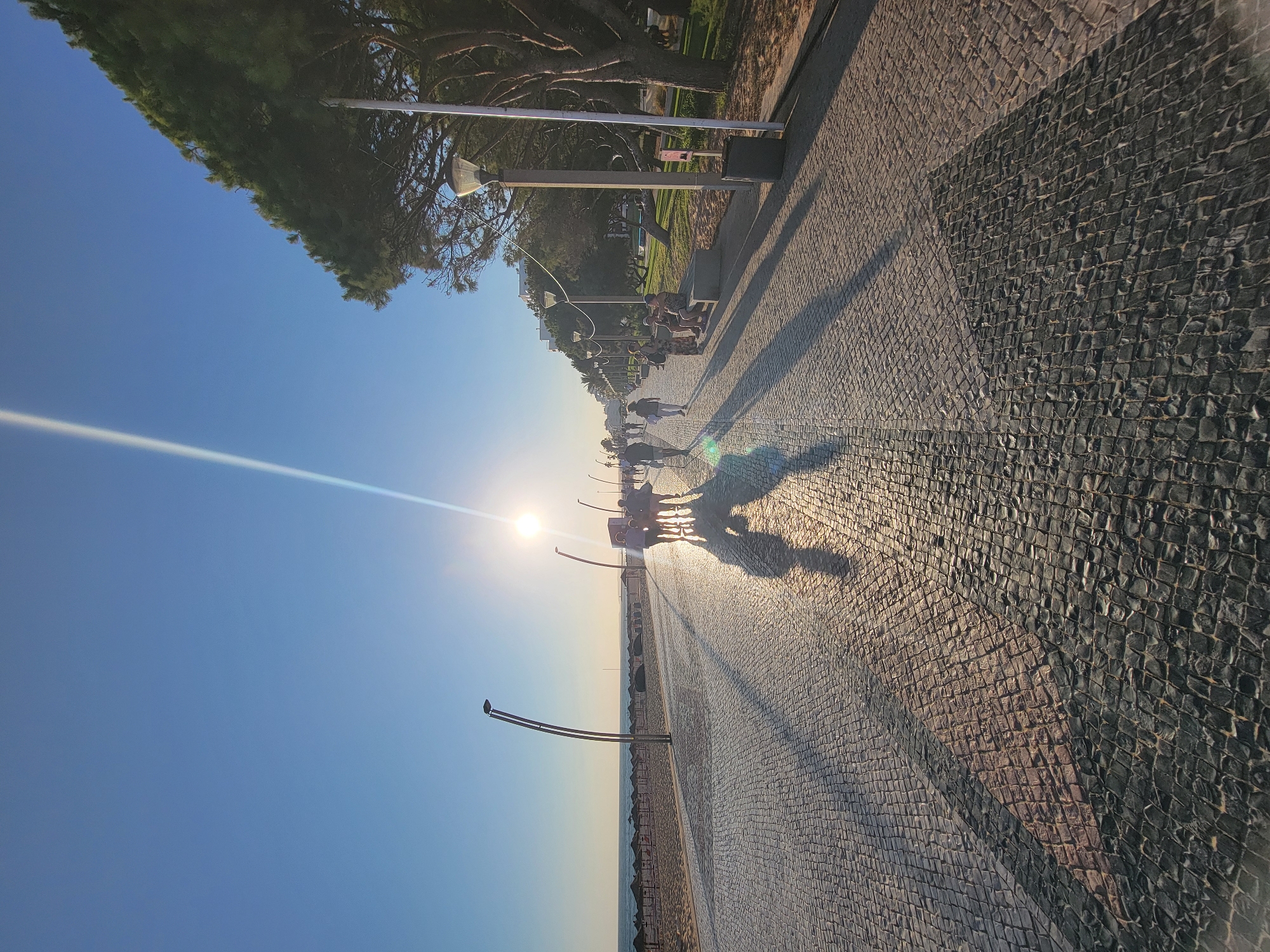 The image captures a sunlit promenade in Quarteira, Portugal, leading towards the ocean. The ground is paved with cobblestones that reflect the bright sunlight, creating long shadows. A group of people are walking along the promenade, casting their own shadows. To the left, there is a sandy beach with umbrellas, and beyond that, the calm ocean under a clear blue sky. On the right side of the promenade, there are trees, lampposts, and some seating areas with people relaxing. In the distance, buildings are visible. The sun is high in the sky, creating a lens flare that streaks across the image.