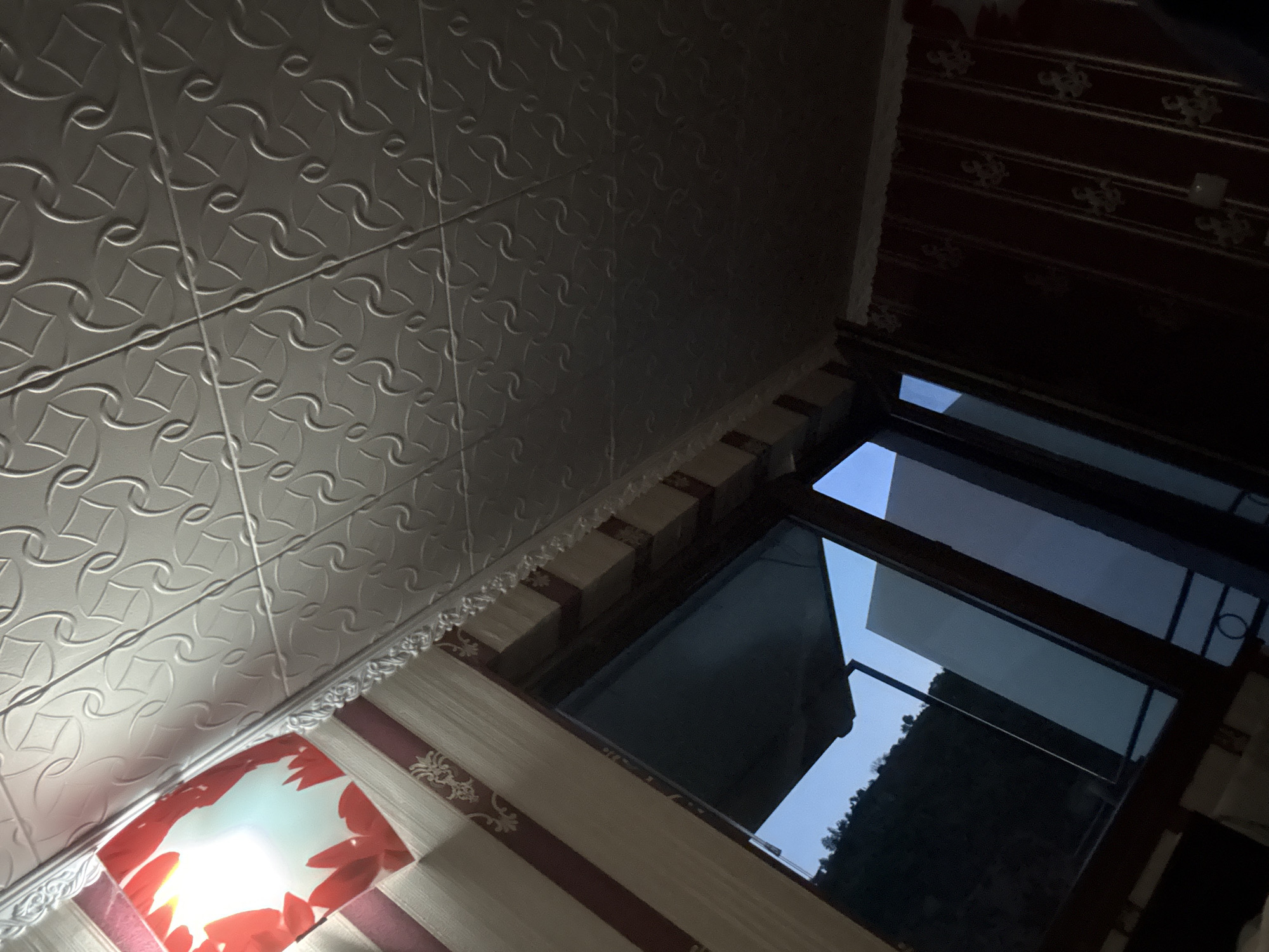 The image shows an interior view from a room with a ceiling that has a textured pattern. There is a light fixture with red floral accents near the top left of the frame. A window occupies a significant portion of the image, revealing a nighttime or dusk view of a hillside with trees against a blue sky. The room's walls are decorated with striped wallpaper featuring a subtle pattern. The overall impression is that of a residential or hotel room in Redina, Bulgaria, at the end of the day. No people or specific actions are visible.