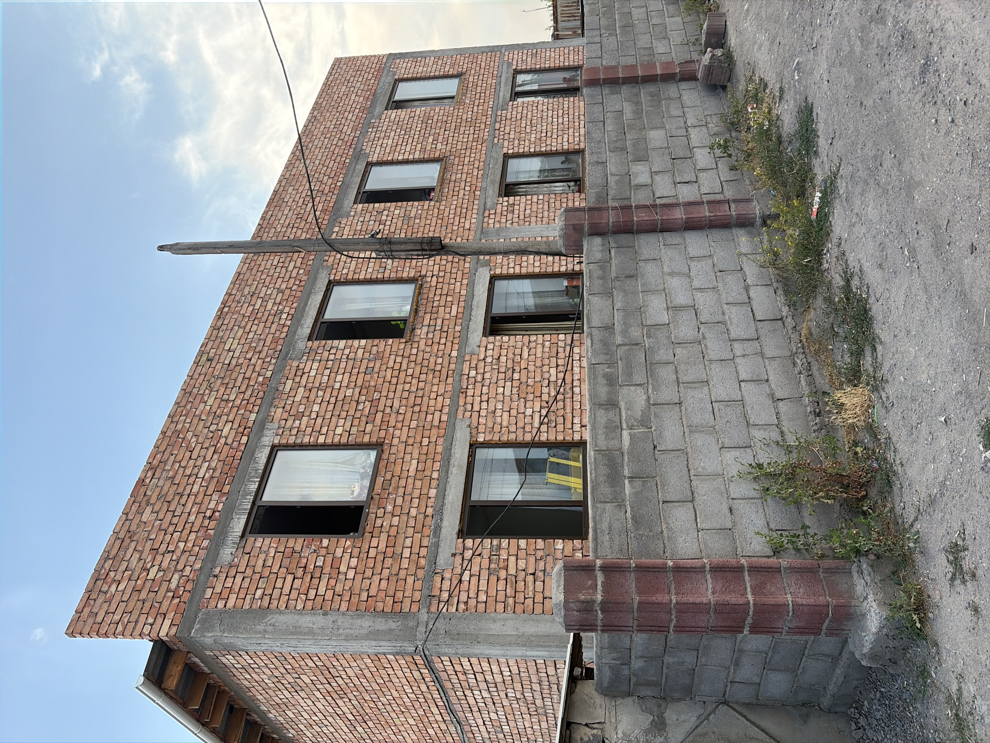 The image shows a brick building under construction in Kant, Kyrgyzstan. The building has multiple stories with many windows. A wooden beam extends horizontally from the building, supported by wires. To the right of the brick building, there is a wall constructed from gray bricks. The ground in front of the wall is covered with interlocking gray pavers and some gravel. Green vegetation and some debris are growing along the base of the gray brick wall. The sky above is mostly clear with some wispy clouds. The lighting suggests it is daytime, likely late afternoon or early evening due to the soft light. No people are visible in the image. The overall scene depicts an urban environment with ongoing construction.