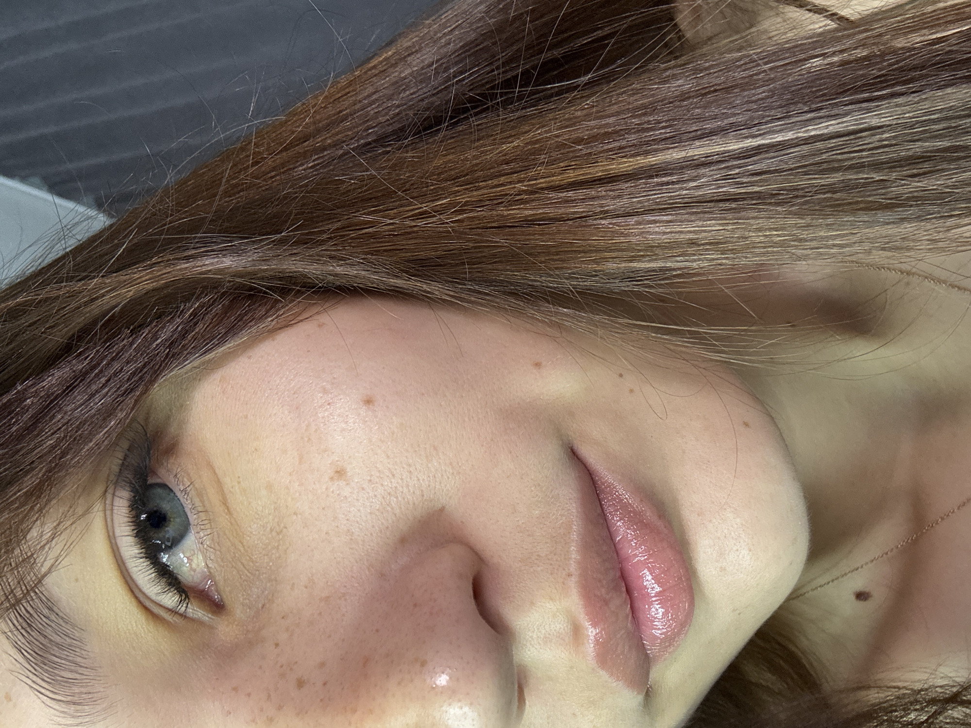 The image is a close-up, horizontally oriented, portrait-style photograph of a person's face, focusing on the eye, nose, and lips. The subject has light skin with scattered freckles across the cheeks and nose. Their eye is visible, with a blue iris and long eyelashes. The lips are painted with a glossy, pink lipstick, appearing full and slightly parted. Brown hair frames the face, with some strands catching the light. The background is out of focus, showing what appears to be an indoor setting with a gray, textured surface behind the subject's head and a lighter surface to the left.
There is no specific activity or event depicted; the image seems to be a posed portrait or a selfie. The lighting suggests it could be daytime, possibly indoors near a window or with artificial lighting. The overall impression is one of a casual, personal photograph. There is no visible text in the image. The location context provided is Novokuznetsk, Russia, but there are no specific visual cues in the image to confirm this.