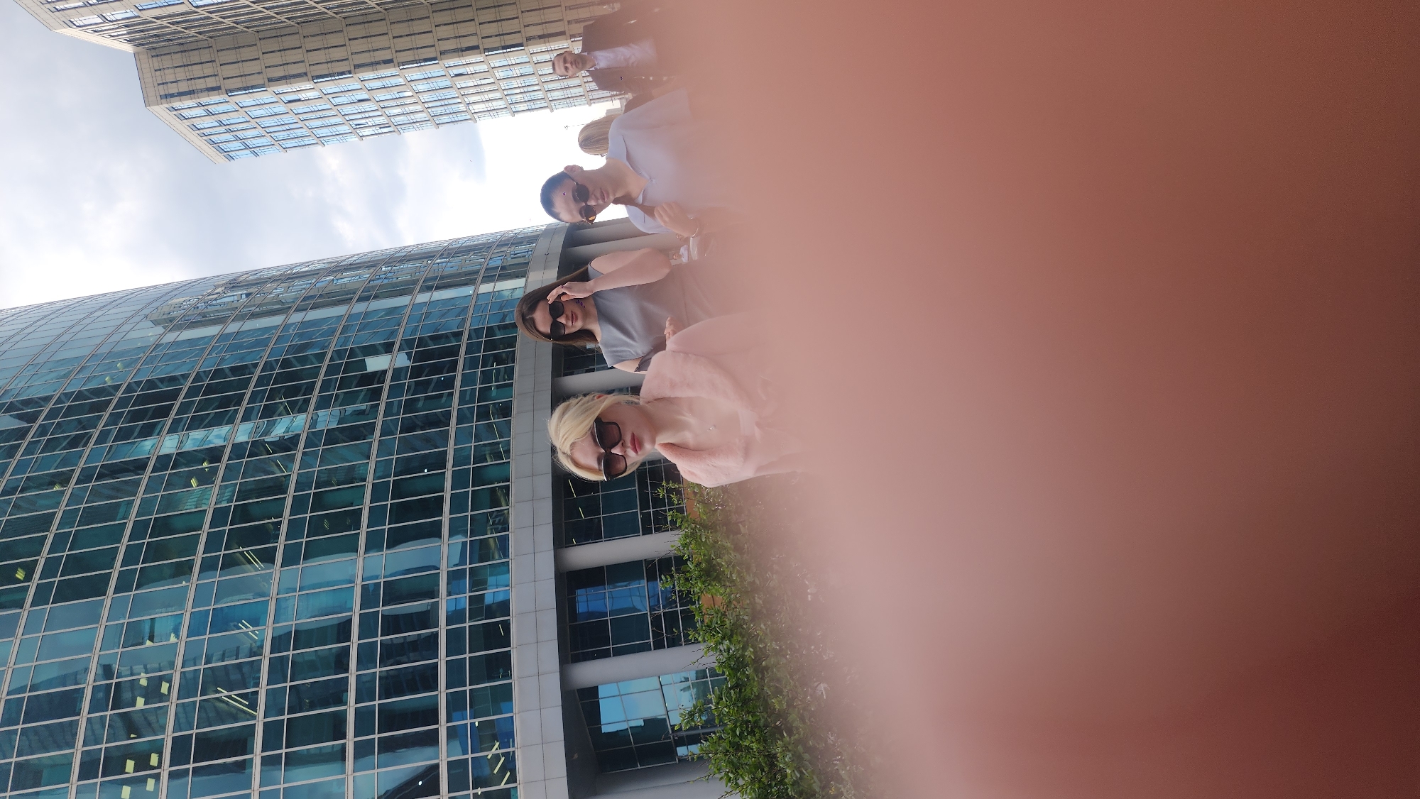 Three individuals stand in front of the Moscow International Business Center (Moscow City), partially obscured by a blurry foreground. The woman in the foreground has blonde hair and wears sunglasses and a pink jacket. Another woman behind her wears sunglasses and a grey shirt. A man stands behind them wearing sunglasses and a light blue shirt. In the background, there are modern glass skyscrapers.