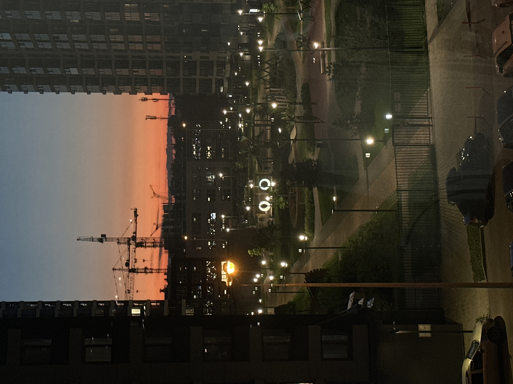 The image presents an urban landscape at dusk or twilight, captured from a high vantage point overlooking a residential development in Perm, Russia. The sky above transitions from a gentle blue at the zenith to a vivid orange and pink at the horizon, signaling a recent sunset.
Tall, modern apartment buildings line the left side of the frame, their numerous windows creating a grid pattern. In the mid-ground, several construction tower cranes stand silhouetted against the colorful sky, indicating ongoing development. Below and around these structures, a network of pedestrian pathways winds through a park or courtyard area, illuminated by numerous glowing streetlights. These lights cast warm glows on green spaces, benches, and what appears to be a playground, featuring a distinct, circular illuminated structure. Other buildings with illuminated windows are visible in the background.
On the right side, a paved area functions as a parking lot, where several cars are visible, their forms indistinct in the low light. A dark metal fence separates this parking zone from the central park area. No people are visible, contributing to a serene atmosphere despite the urban setting. The artificial light from the streetlights and building windows stands in contrast to the fading natural light of the sky. Small, white rectangular signs with unreadable text are barely discernible on dark utility boxes or building facades near the parked vehicles.