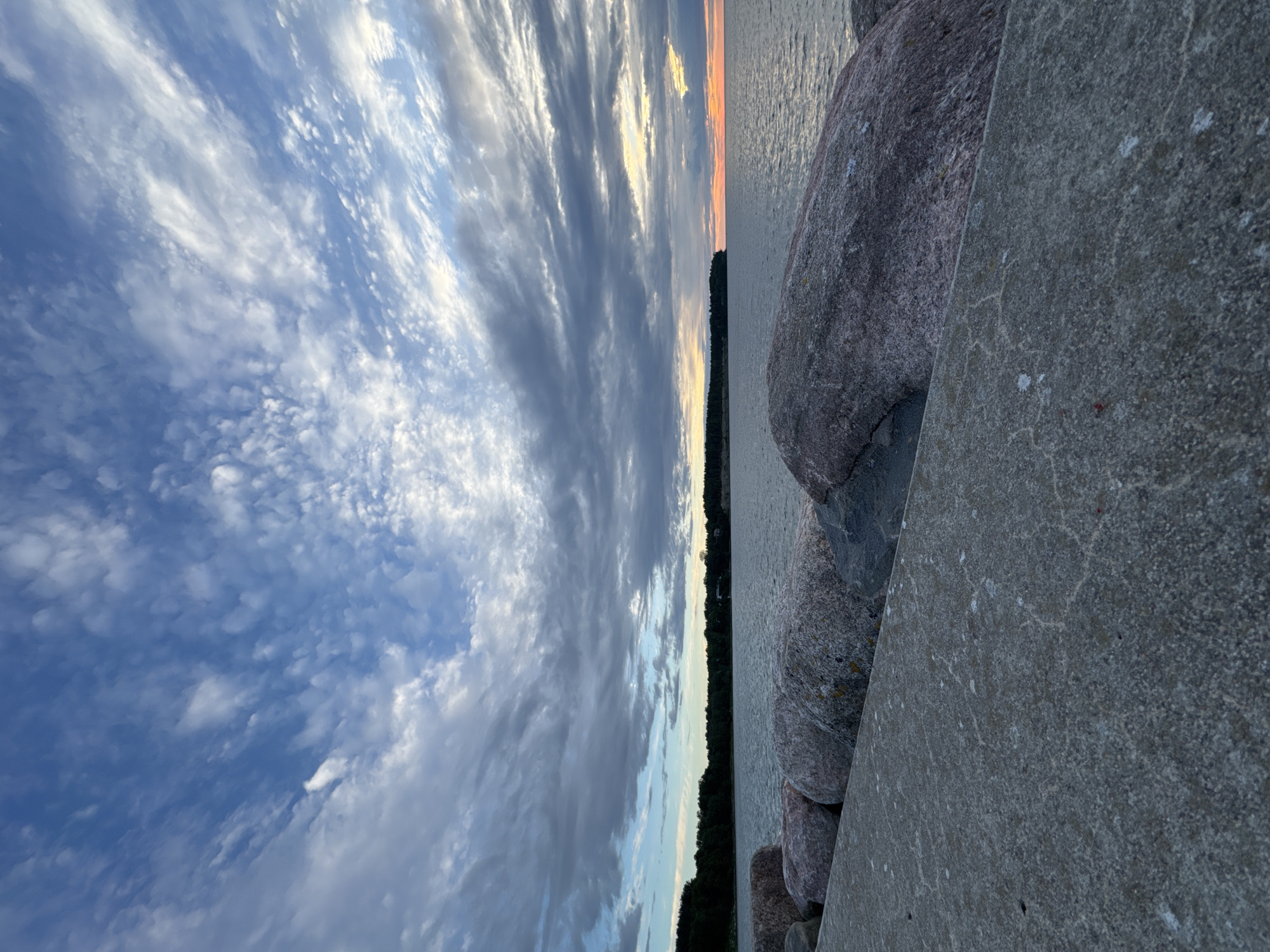 The image captures a view of the Baltic Sea from a concrete pier dotted with large, rough-hewn rocks, likely part of a breakwater or coastal fortification. In the distance, a dense line of dark green trees forms the shoreline, suggesting a forested coastal area. The sky above is a dramatic canvas of blues and grays, with wispy white clouds interspersed with larger, more solid formations. Towards the right horizon, the sky is illuminated with soft hues of orange and yellow, indicating either sunrise or sunset. The water is calm, reflecting the colors of the sky, and a gentle ripple suggests a light breeze. The foreground is dominated by the textured surface of the concrete pier and the rounded shapes of the large, pink-tinged rocks, some showing lichen growth. The scene is devoid of any human presence or activity. This scene is consistent with the coastal areas of Tallinn, Estonia, particularly regions like Pirita or Nõmme, known for their breakwaters and forested coastlines along the Baltic Sea.