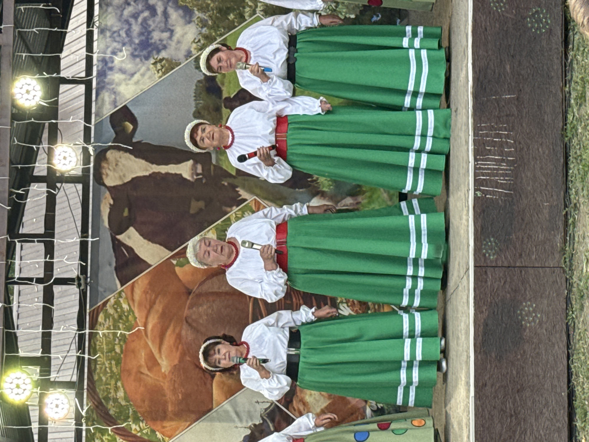 Four women are standing on a stage in front of a backdrop depicting a rural scene with a cow and bread. They are all wearing traditional folk costumes consisting of white blouses with long sleeves, red sashes, and long green skirts with white trim. Each woman is holding a microphone and appears to be singing. The stage is wooden, and there are lights overhead. The event is taking place in the city of Кукуи, Russia.