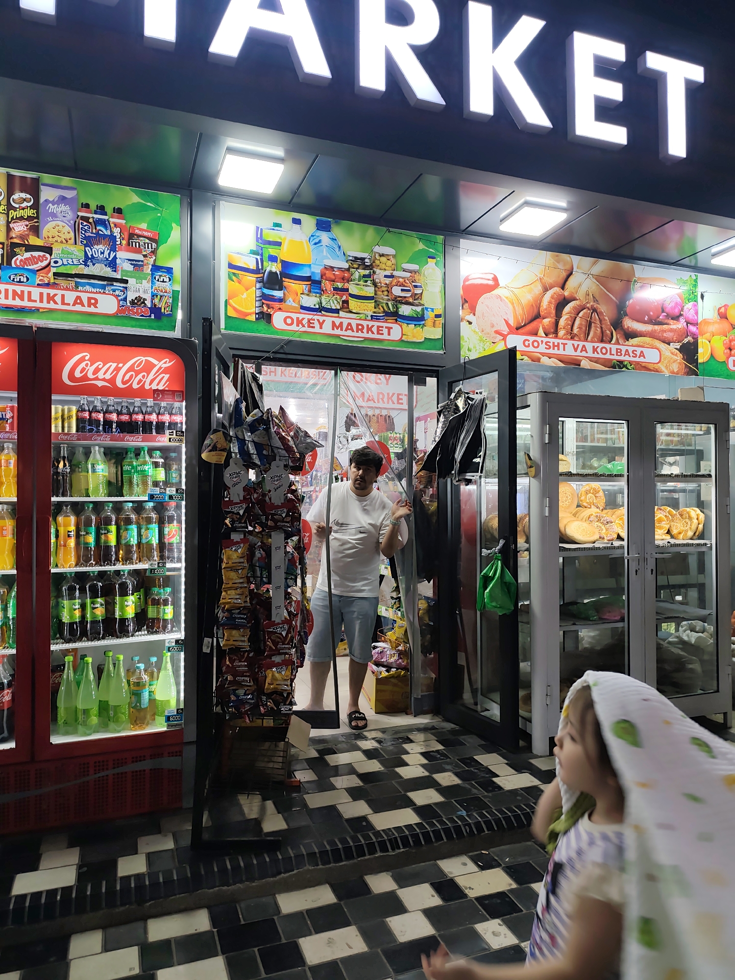 A man stands in the entrance of an "OKEY MARKET" convenience store in Fergana City, Uzbekistan. The store's facade features large illuminated signs, displaying advertisements for snacks and drinks. A red Coca-Cola refrigerator unit is visible on the left, stocked with various beverages. To the right, a display case showcases baked goods, including loaves of bread and pastries. The man, wearing a white t-shirt and blue shorts, is looking towards the right side of the frame. A young child, partially obscured by a white cloth, is walking in the foreground. The floor is tiled in a black and white checkerboard pattern.