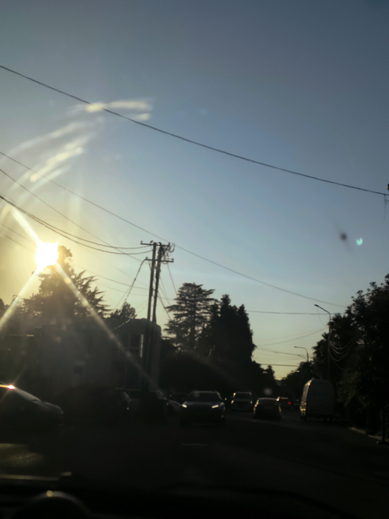 The image captures a view from inside a vehicle looking out at a street scene in Salibauri, Georgia, likely during late afternoon or early evening, given the low angle of the sun and lens flares. The sun, a bright white orb with strong lens flares creating diagonal streaks of light, is visible on the left side of the frame, partially obscured by trees and power lines. Several cars are on the road, appearing as silhouettes against the brighter sky and roadside. A utility pole with multiple wires stretches across the upper portion of the image. Trees line the sides of the road, with their silhouettes dark against the sky. The road surface is dark asphalt. The sky is a gradient of light blue, fading to a lighter hue near the horizon. There are no discernible people visible in the image. The overall impression is of a typical urban street during a sunset, with traffic and infrastructure elements clearly visible.
