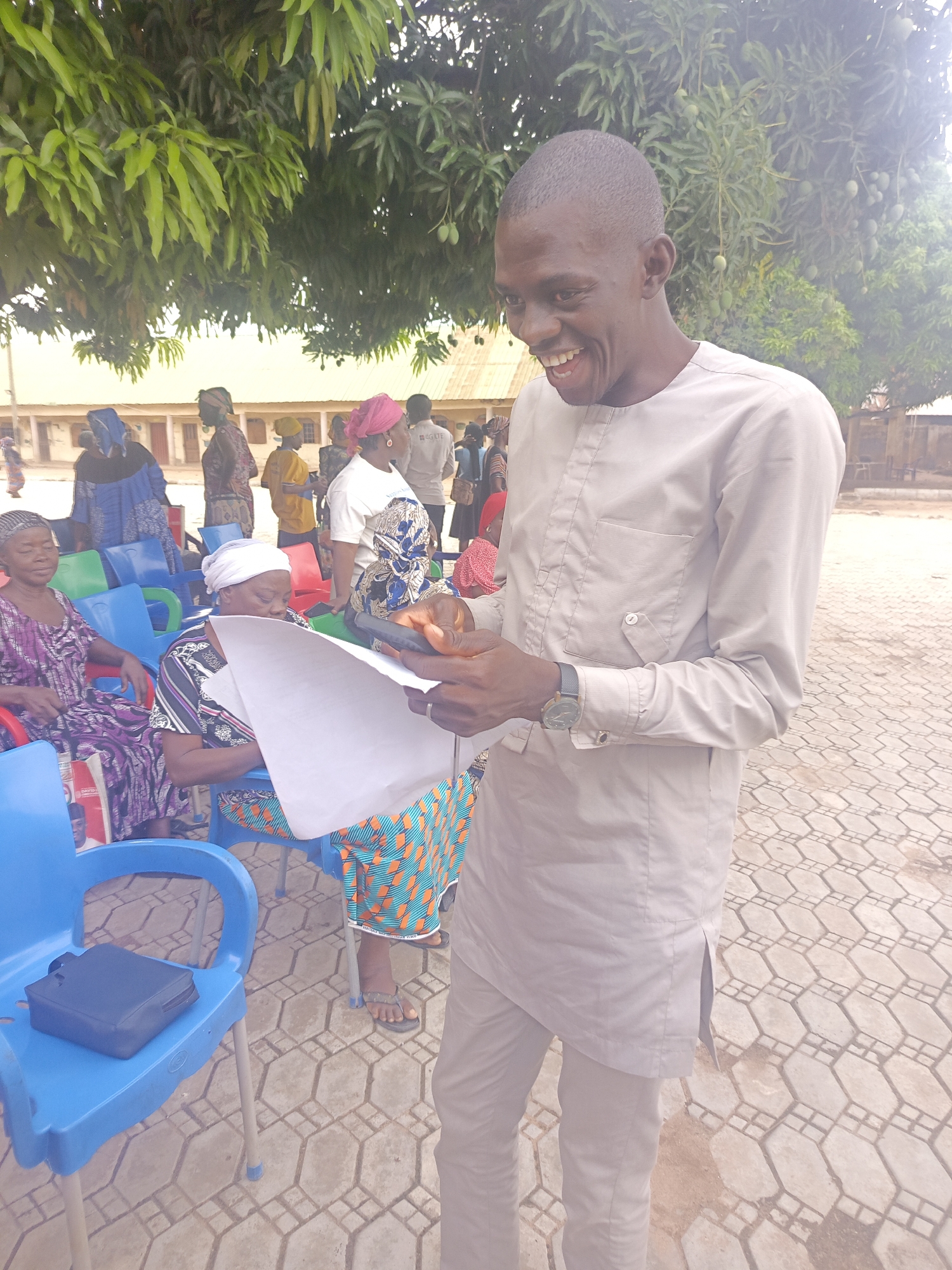 The image depicts a man in a light grey traditional Nigerian outfit, smiling while looking at a white sheet of paper. He is outdoors on a paved surface. Behind him, a group of people, mostly women in colorful traditional attire, are seated on blue and green plastic chairs. A black purse sits on one of the chairs. Overhanging the scene is a large leafy tree. The setting is likely a public space in Akwanga, Nigeria.