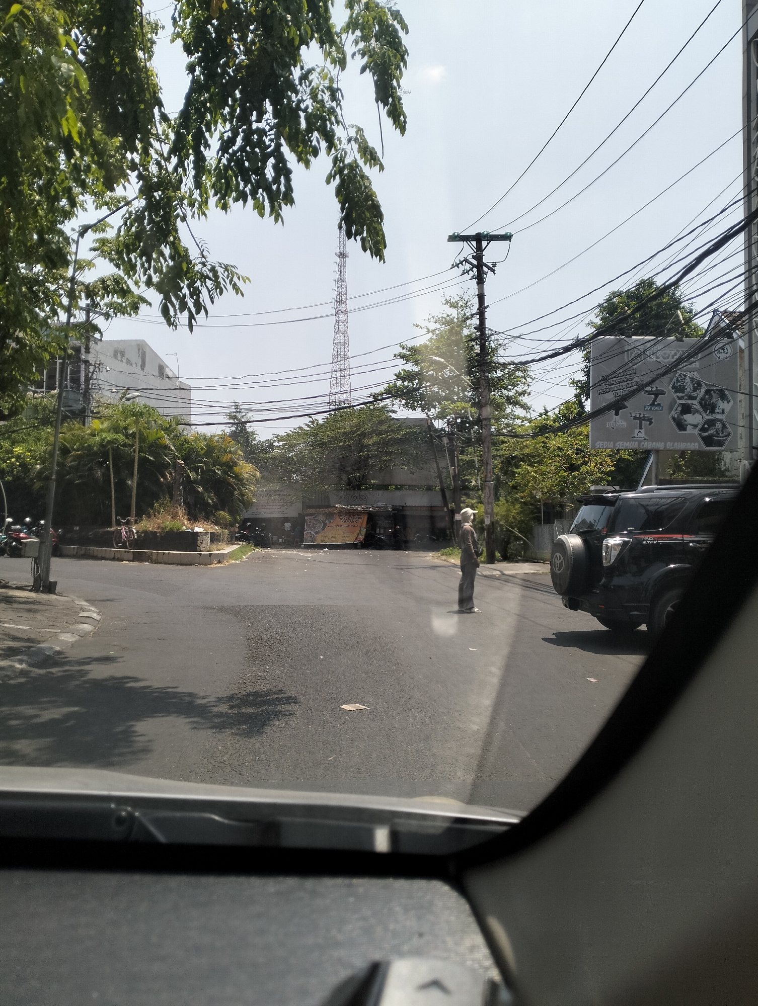 A person wearing a light-colored hat and pants stands on the side of a road in Semarang, Indonesia, during the daytime. The person is looking towards the right side of the frame. A black SUV is parked on the right side of the road, facing left. In the background, there is a tall communications tower, trees, buildings, and power lines. The road is asphalt. The image is taken from inside a vehicle, looking out through the windshield, which is partially visible in the foreground. The scene appears to be an intersection.
