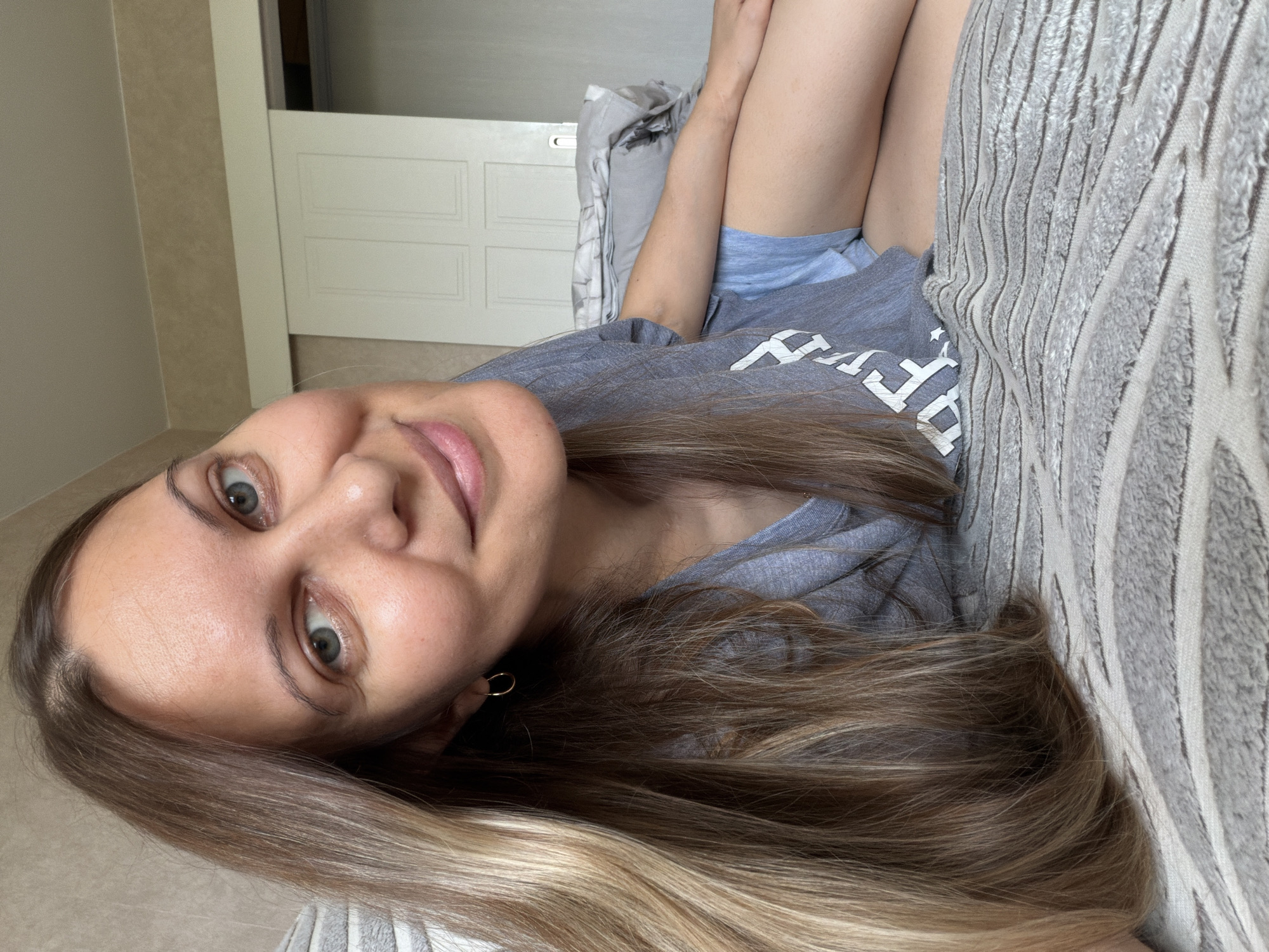 A woman with long, blonde-brown hair is lying on a bed, looking at the camera with a smile. She is wearing a grey t-shirt and blue shorts. The bed is covered with a grey and white striped blanket. In the background, there is a sliding closet door. The room appears to be a bedroom. The location in Izhevsk, Russia is not discernible from the image.