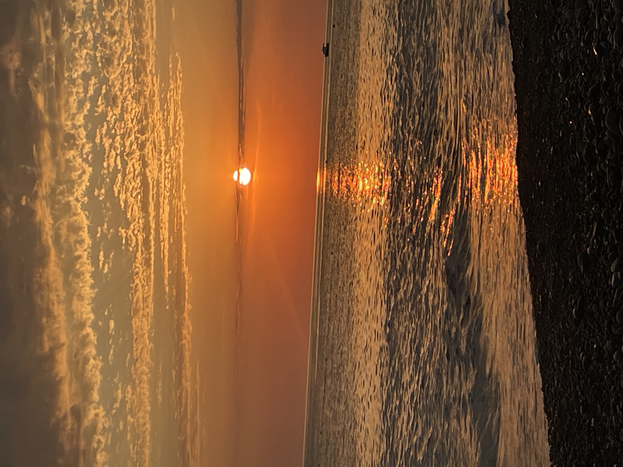 A vibrant sunset paints the sky in hues of orange and gold, with wispy clouds stretching across the horizon. The sun, a bright orb, peeks through a thin layer of clouds, casting a shimmering reflection on the calm, rippling water of the ocean. A small boat is visible in the distance, sailing across the tranquil sea. The foreground is occupied by a dark, pebbly beach, with the stones creating a textured foreground. The scene captures the serene beauty of a coastal sunset.
