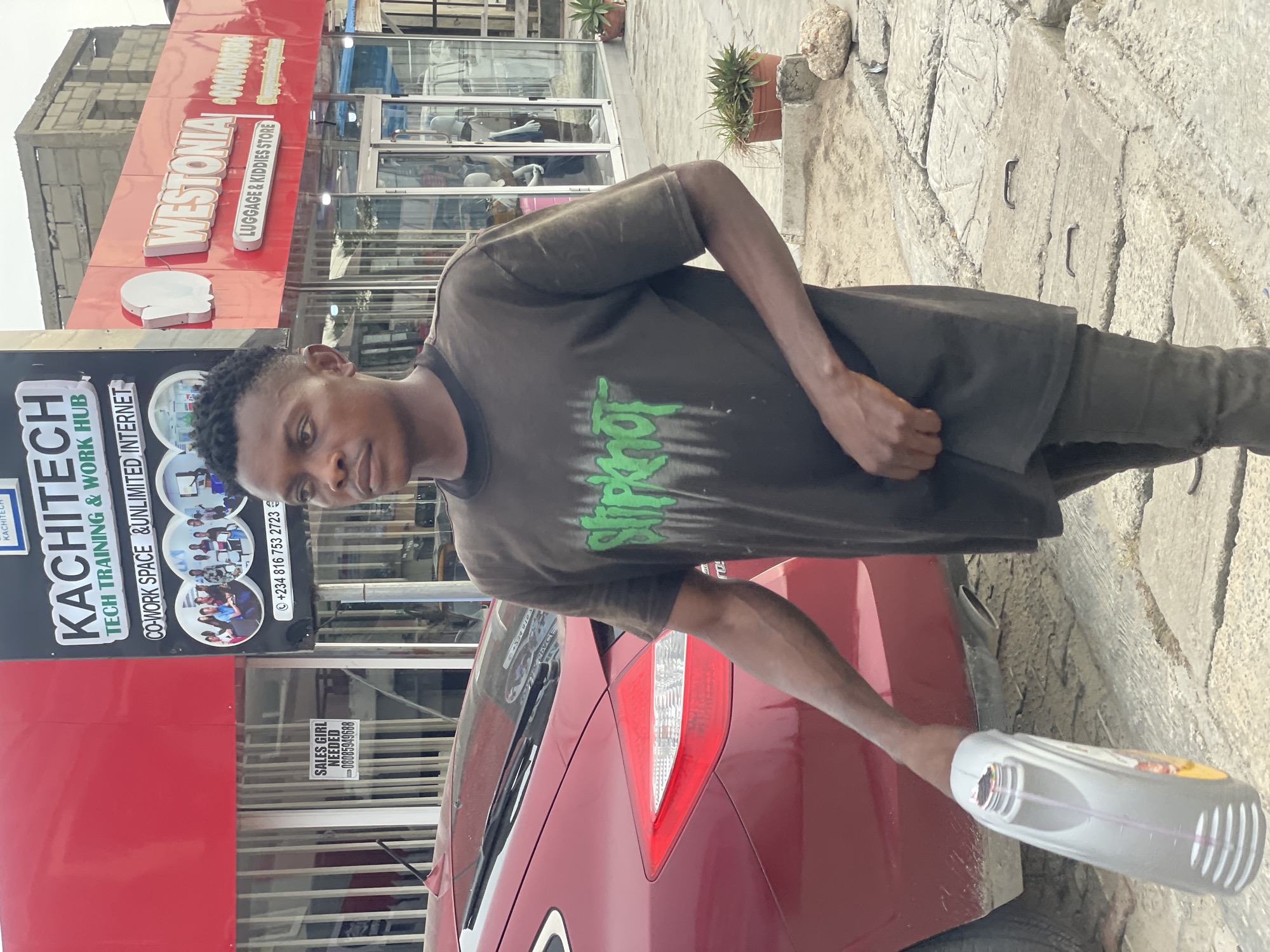 An individual is standing by the rear of a red car, holding a partially empty oil bottle. He wears a dark-colored t-shirt with green writing and dark trousers. In the background is a building with visible signs, including "KACHITECH TECH TRAINING & WORK HUB" with the phone number "+234 816 753 2723" and "WESTONIA LUGGAGE & KIDDIES STORE" with the phone number "09014098384". Also visible is a small sign on the car stating "SALES GIRL NEEDED - 08085949688". The image was taken in Ado, Nigeria.