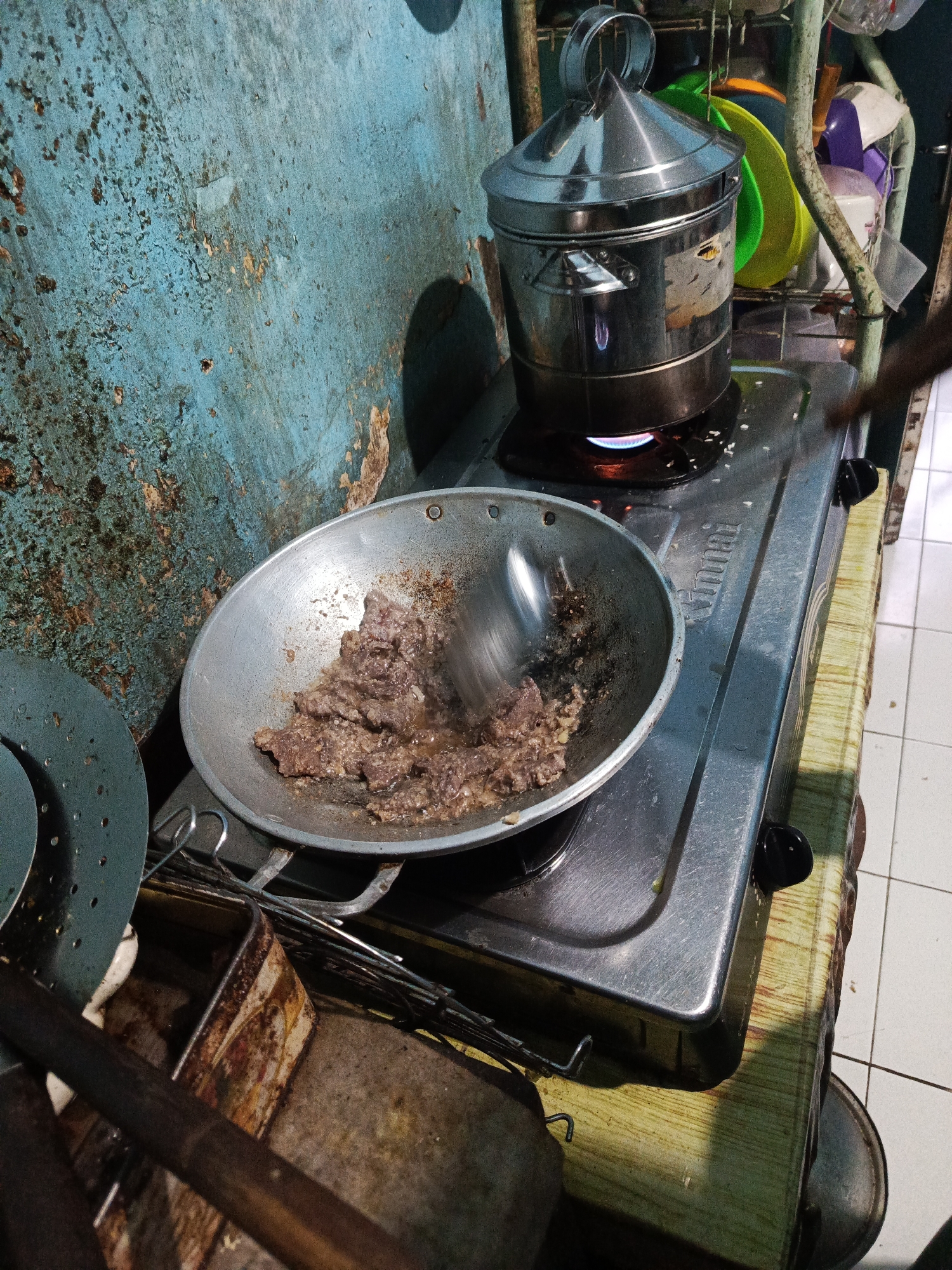 The image depicts a home kitchen scene in Kandangjati Kulon, Indonesia, focusing on a cooking process. In the foreground, a metal wok containing pieces of browned meat is positioned on a gas stove. A spoon is actively stirring the meat, suggesting it is being fried or sautéed. Adjacent to the wok, on the left side of the stove, is a perforated metal disc, possibly a steamer tray or a colander. To the right of the wok, a stainless steel steaming pot with a conical lid is placed on another burner of the same gas stove, with a blue flame visible underneath, indicating it is also in use, likely heating or steaming food.
The background reveals a rustic and weathered blue wall, showing signs of rust and peeling paint. Behind the stove and steaming pot, a metal rack holds various kitchen items, including colorful plates and containers, along with a plastic bottle. The overall environment suggests a functional, albeit aged, cooking area. The floor appears to be tiled. The image captures a moment of everyday life, highlighting the preparation of food in a domestic setting.