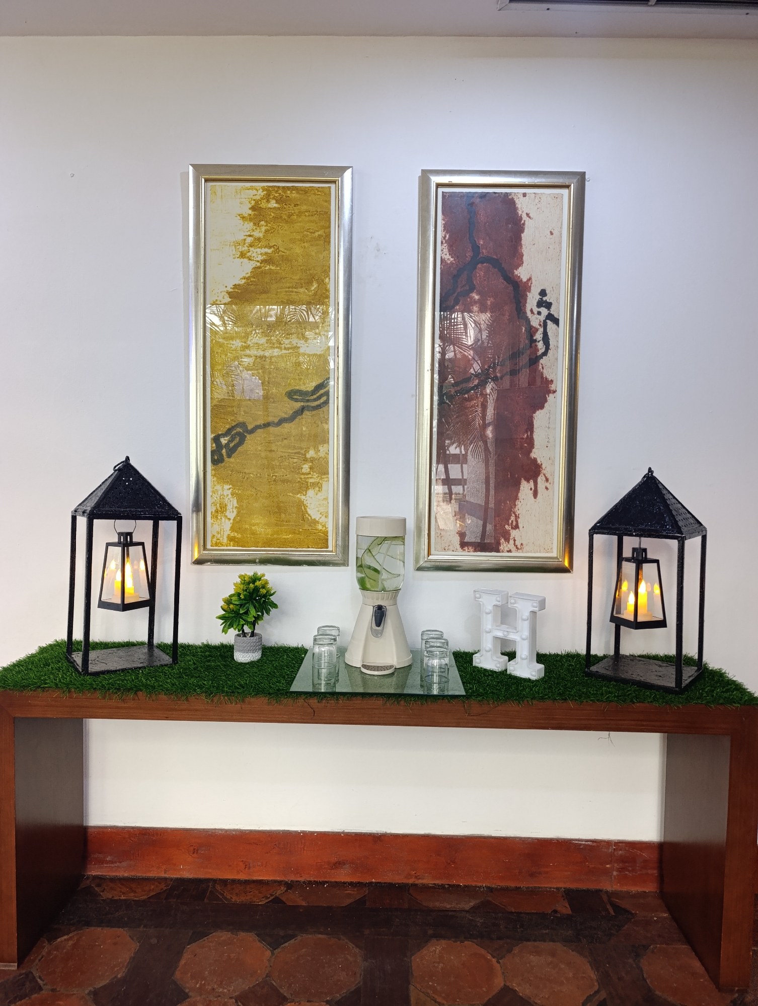 The image shows an interior setting, seemingly a foyer or hallway, in an unknown location in an unknown city.
Against a white wall, two framed abstract art pieces hang, each in a silver frame. The left artwork features a yellow and white color scheme with black lines, while the right piece has a red, white, and black motif.
A dark-stained wooden console table is positioned below the artworks. The table top is covered with a synthetic green turf. On the turf, from left to right, are: a black metal lantern with a lit candle inside, a small potted green plant, two clear drinking glasses, a clear drink dispenser filled with cucumber-infused water, another two clear drinking glasses, and a white block letter "H" decoration, ending with another black metal lantern matching the one on the left.
The console table has a lower shelf, also dark-stained wood. The floor is tiled with octagonal tiles in shades of brown. The overall setting appears staged, possibly for a hotel lobby or a decorative display. There are no people visible in the image.
