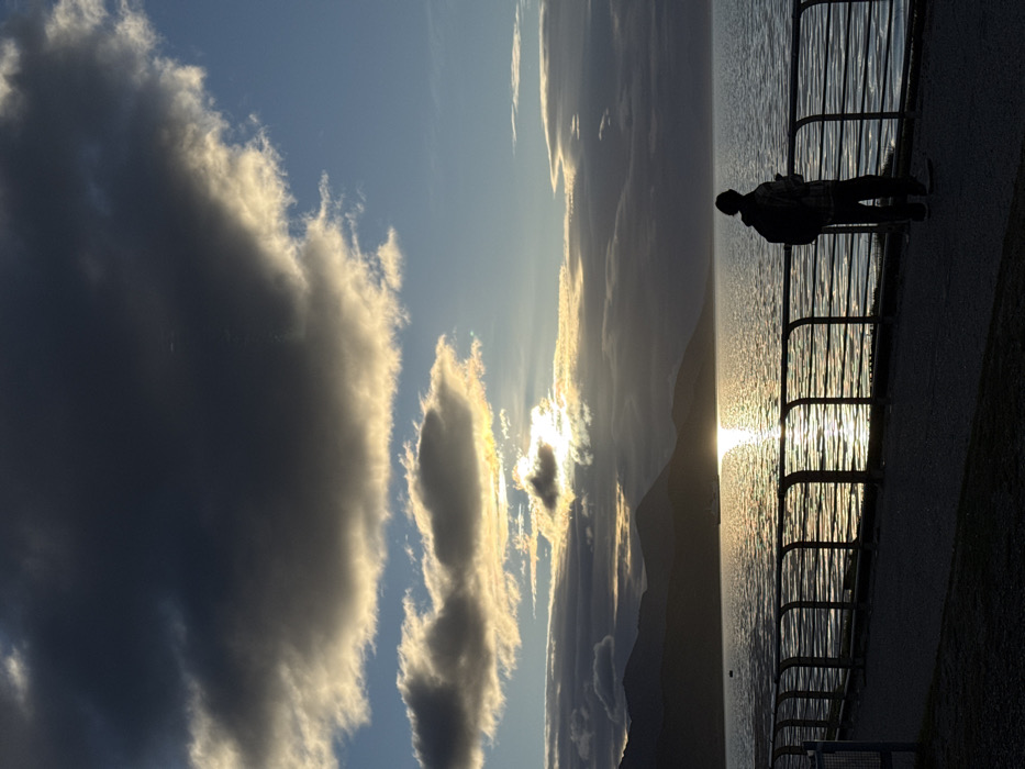 The image displays a waterfront scene in Ushuaia, Argentina, during what appears to be late afternoon or early evening. The sky is partially covered by dark grey and illuminated clouds, with visible patches of blue. The sun, partially obscured by clouds, is low on the horizon, casting a bright golden reflection across a large body of water, likely the Beagle Channel. A range of dark, silhouetted mountains forms the distant horizon. In the foreground, a paved promenade runs parallel to the water, separated by a dark metal railing with horizontal bars. A single individual stands in silhouette on the promenade, positioned slightly right of center, facing the water. The person wears a backpack and holds a small object, possibly a mobile device, in their right hand. A small, light-colored vessel is visible on the water within the sun's reflection, and a dark object, likely a buoy, floats closer to the left shore. No other individuals or direct interactions are depicted.