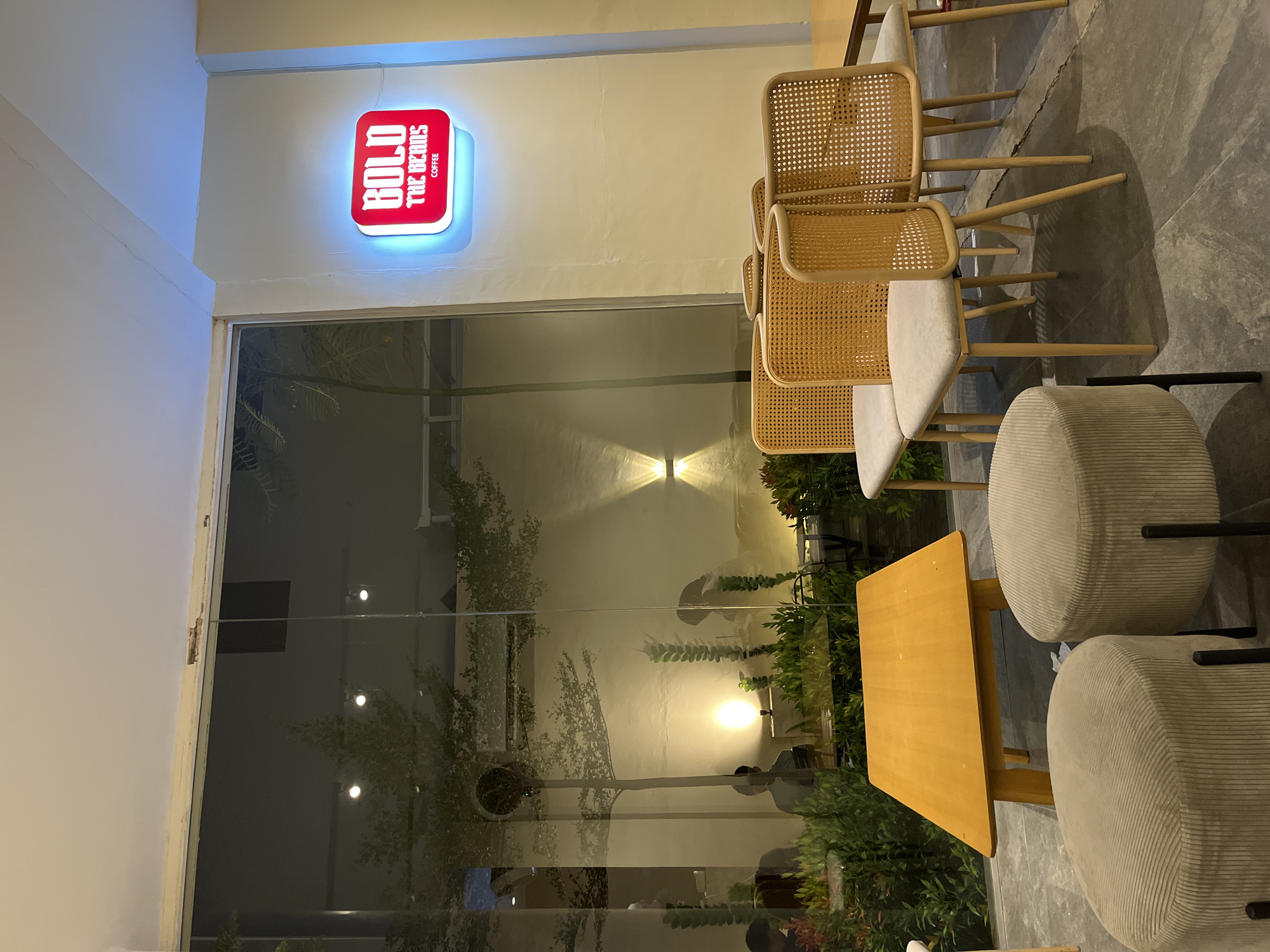 The image depicts the interior of a coffee shop in Lhokseumawe, Indonesia. A red and white illuminated sign reading "BOLD THE BEANS COFFEE" is mounted on a cream-colored wall to the right. In the foreground, two beige, cylindrical ottomans with ribbed textures sit on a dark gray floor. Behind them is a light yellow, trapezoidal coffee table. Several chairs with light gray upholstered seats and woven rattan backs are arranged in a cluster. A large glass window occupies the left side of the image, offering a view of the exterior. Through the window, trees with sparse foliage are visible, illuminated by warm outdoor lighting. Silhouettes of people can be seen inside the coffee shop through the glass, some seated at tables. The overall ambiance is modern and relaxed, with a focus on natural materials and ambient lighting.