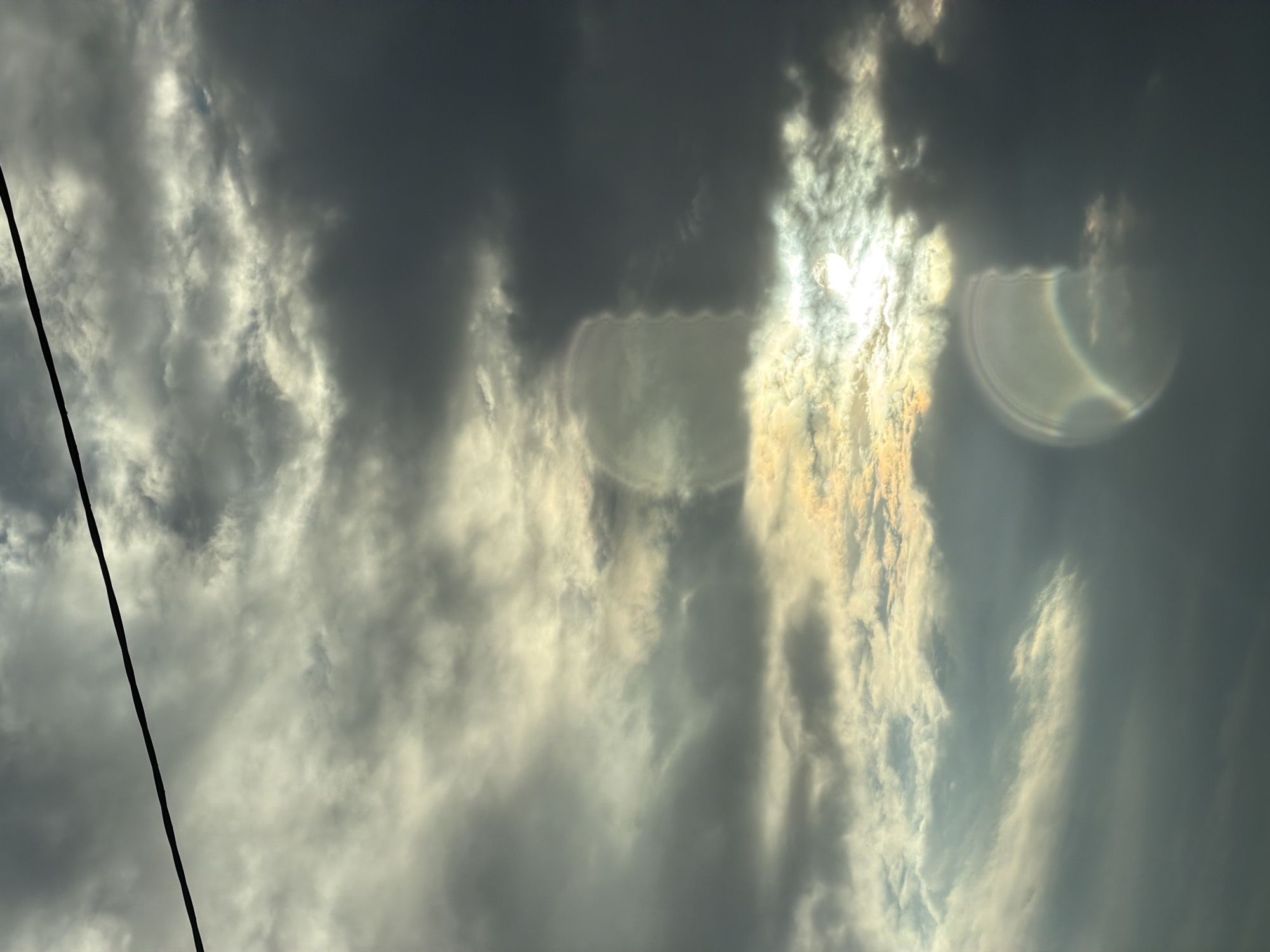 A dramatic, overcast sky is captured from below, with dark, heavy clouds dominating the frame. A thin, black wire is stretched diagonally across the upper portion of the image, from the top left to the middle right. Patches of sunlight are breaking through the clouds, creating bright, luminous areas. There are two distinct circular lens flares visible in the sky, one in the upper middle and another in the lower right, both exhibiting a prismatic rainbow effect. The overall impression is one of a moody and dynamic sky.