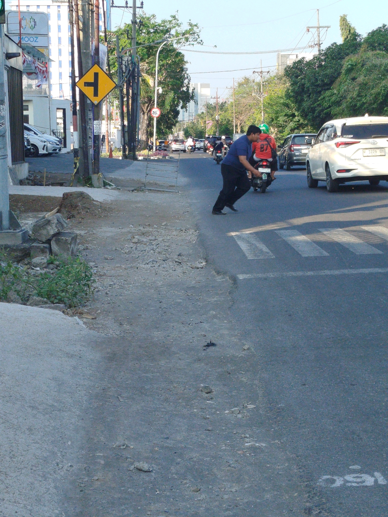A man in a blue shirt and black pants is pushing a motorcycle with a rider in a red shirt with the number 87 on the back, in a street with a pedestrian crossing in Surabaya, Indonesia. The man is on the left side of the motorcycle, pushing it forward. The rider is seated on the motorcycle, looking forward. There are cars and other motorcycles on the road. The street is paved with asphalt and has white lines marking the pedestrian crossing. There are trees and buildings on the sides of the street. The sky is clear and blue. A yellow traffic sign with a black arrow indicating a T-intersection is visible on the left side of the image.