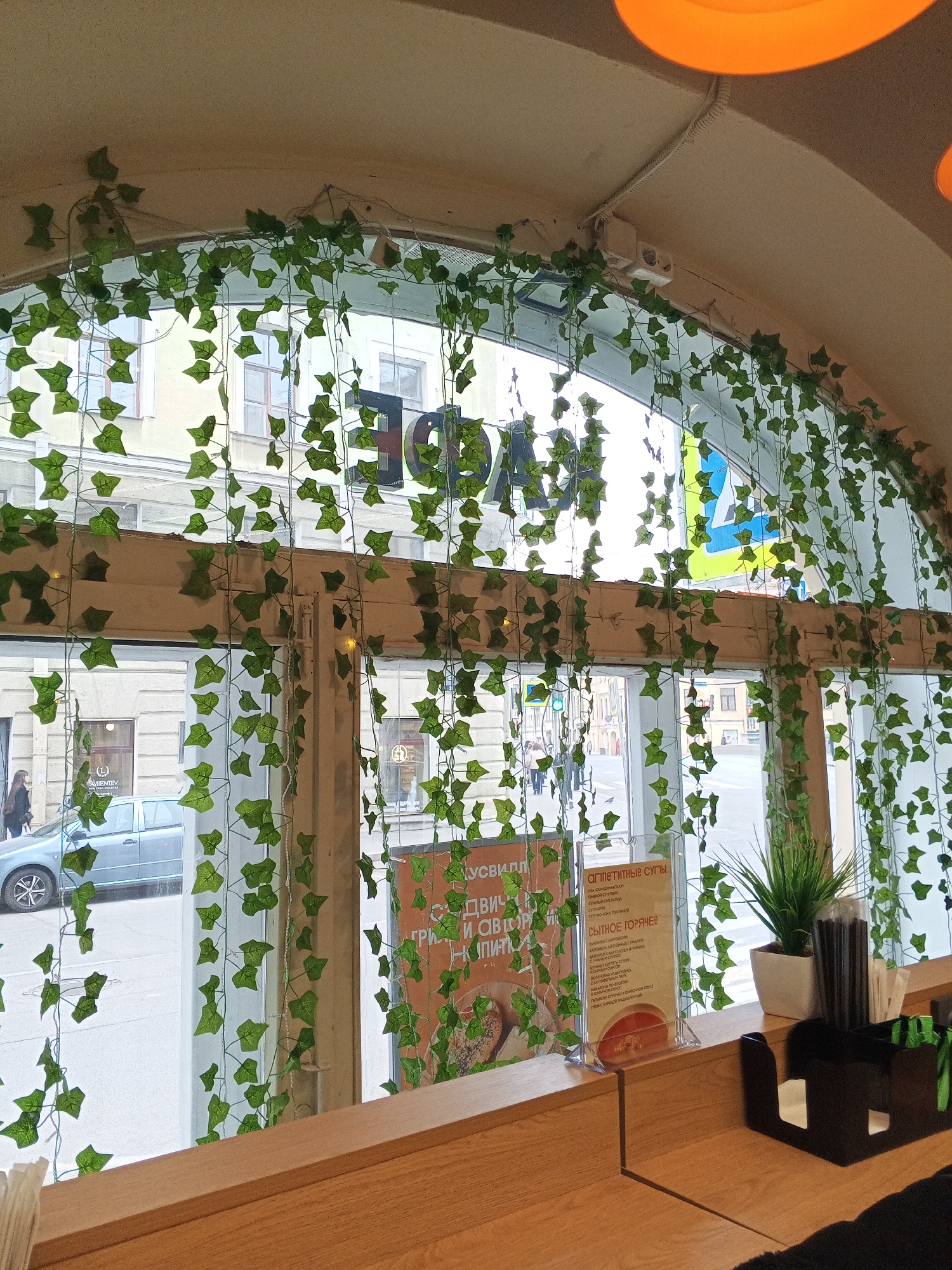 Here is a description of the image:
**Setting:**
* The image appears to be taken from inside a cafe or restaurant in Saint Petersburg, Russia. The view is through a large arched window, decorated with artificial ivy.
* Outside the window, there's a view of a street with buildings on either side. A light-blue car is parked on the street.
**Objects & Items:**
* The interior setting of the cafe shows a wooden counter or bar.
* Menu card with Russian text on it. It lists "Appetizing Soups" and "Hearty Hot Dishes".
* A small potted plant on the counter.
* A container with straws, and a black holder containing other items.
**People:**
* There are people visible on the street through the window, giving a sense of the street's activity.
**Text:**
* The window has partially obscured signs in Russian. The sign says "Face".
* The menu lists a range of soups and hot meals with Russian names.